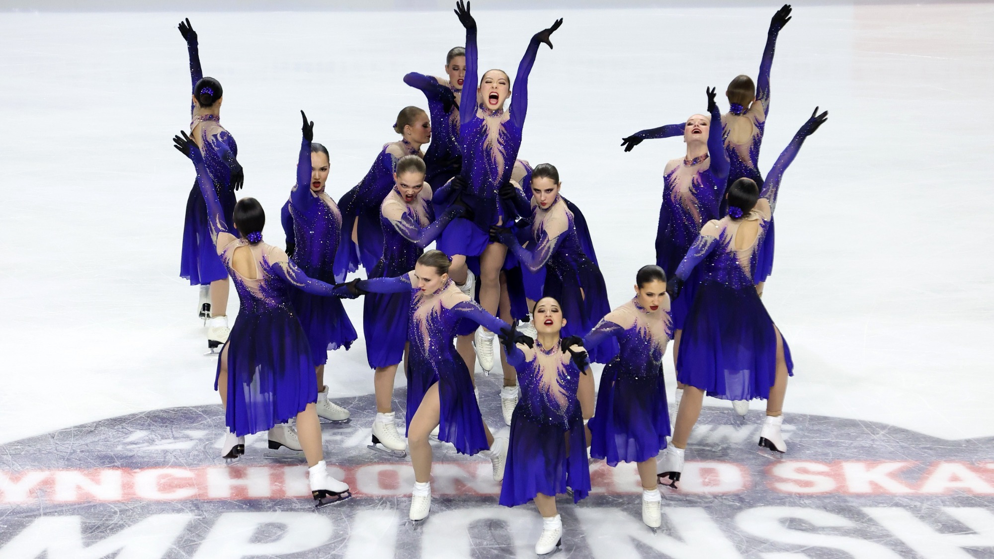 The Haydenettes, in purple dresses that fade to black at the waist and have gems on the top, have fierce expressions. They lift a skater, with other skaters forming a semicircle in front of her.