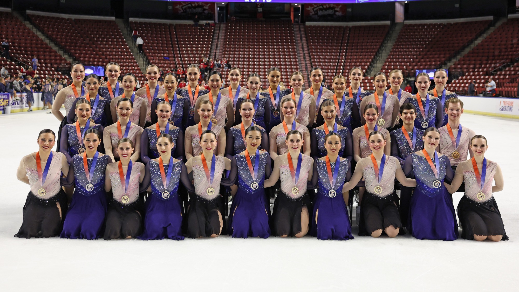 The Haydenettes and Skyliners alternate skaters in a formal World Team photo. Skaters are on the ice in three rows, the last row standing, the middle row sitting, and the closest row kneeling. The Haydenettes wear costumes with light pink tops and black skirts while the Skyliners wear blue costumes with rhinestones on the top.