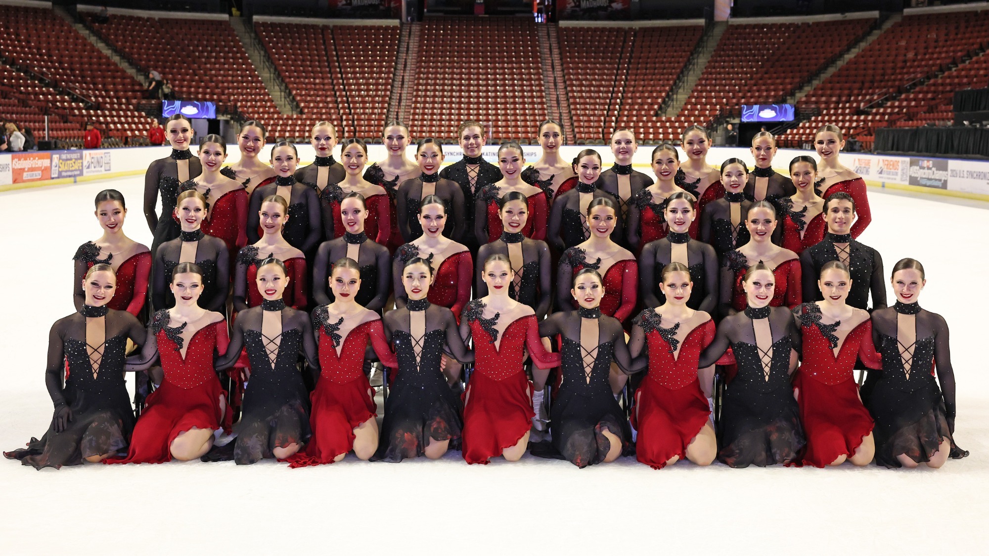 The formal Junior World Team photo. Teams Elite junior skaters, in black costumes with rhinestones on the top, alternate with Skyliners junior skaters, who wear red dresses with a deep V neck, rhinestones on the top and a black detail on the left shoulder. They pose in four rows, the first row kneeling, the second sitting and the last two standing. 
