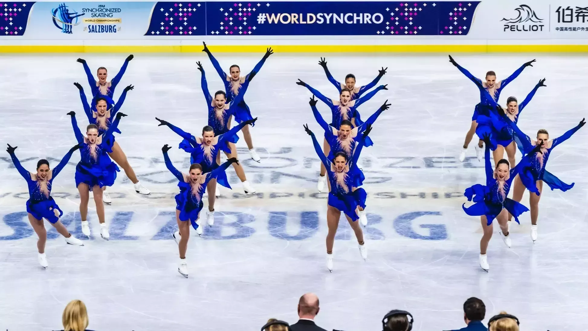 The Haydenettes stand on the ice in four vertical lines of four skaters. Each athlete has their arms extended upward with a look of determination on their face. The athletes are wearing a long sleeve purple skating costume with purple and pink detailing on the front resembling flames and a long purple and black ombre skirt.