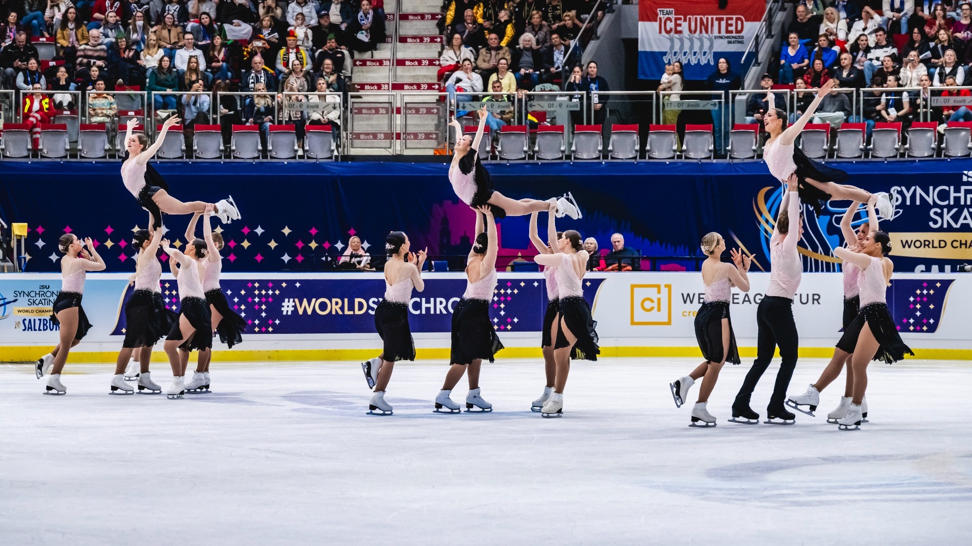 The Haydenettes perform three lifts in unison with one skater being held up by their teammates. They are wearing coordinating long sleeve skating costumes with a light pink top and black skirt. 