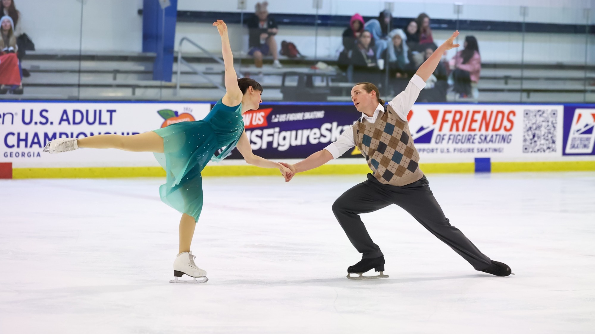Jodi Beggs and Keiffer Hubbell perform holding hands with both skaters in a spiral position. Jodi (left) is a woman wearing a ombre teal dress and Keiffer (right) is a man wearing a tan sweater vest over a white button down shirt and black pants. 