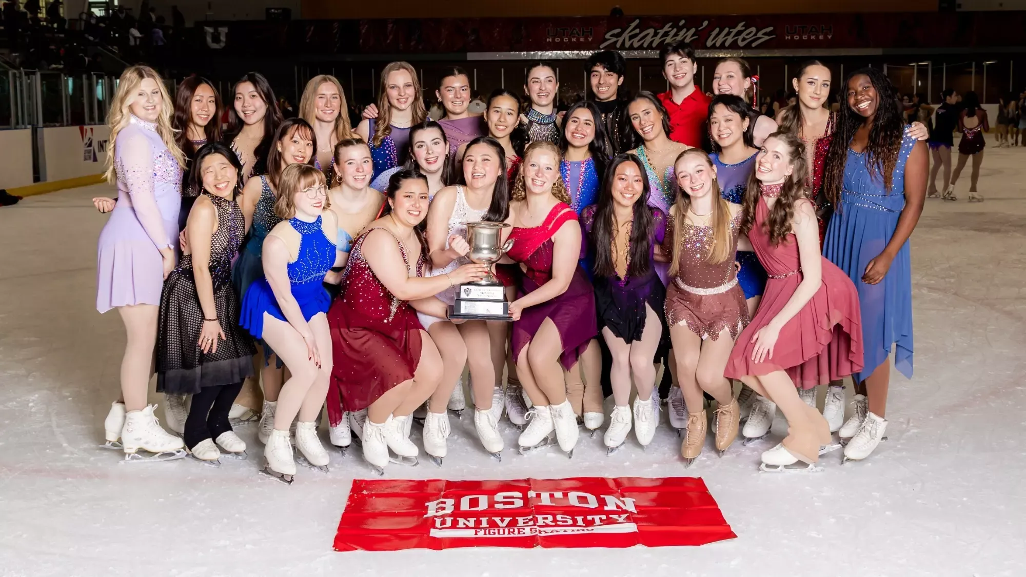 Boston University poses with their large silver National Intercollegiate Final trophy. Skaters are standing in three lines wearing skating costumes of varying styles and colors. In front of them on the ground is a red flag with the words 