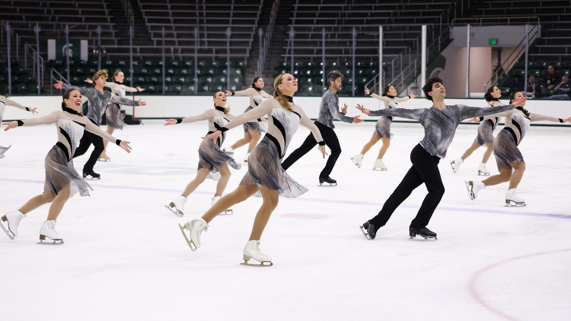 The Skating Club of Boston's Forte of Boston senior TOI team skates in unison with their arms extended wearing coordinating black, white and grey long sleeve costumes. 
