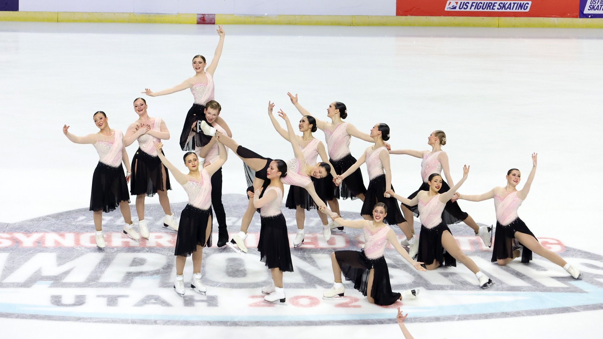 The Haydenettes strike a pose at center ice with one skater behind held up on the shoulders of her teammate. All athletes have their arms extended outward. The skaters are all wearing matching long skating costumes with a light pink long sleeve top and a black skirt. One male skater is wearing a coordinating top with black pants. 