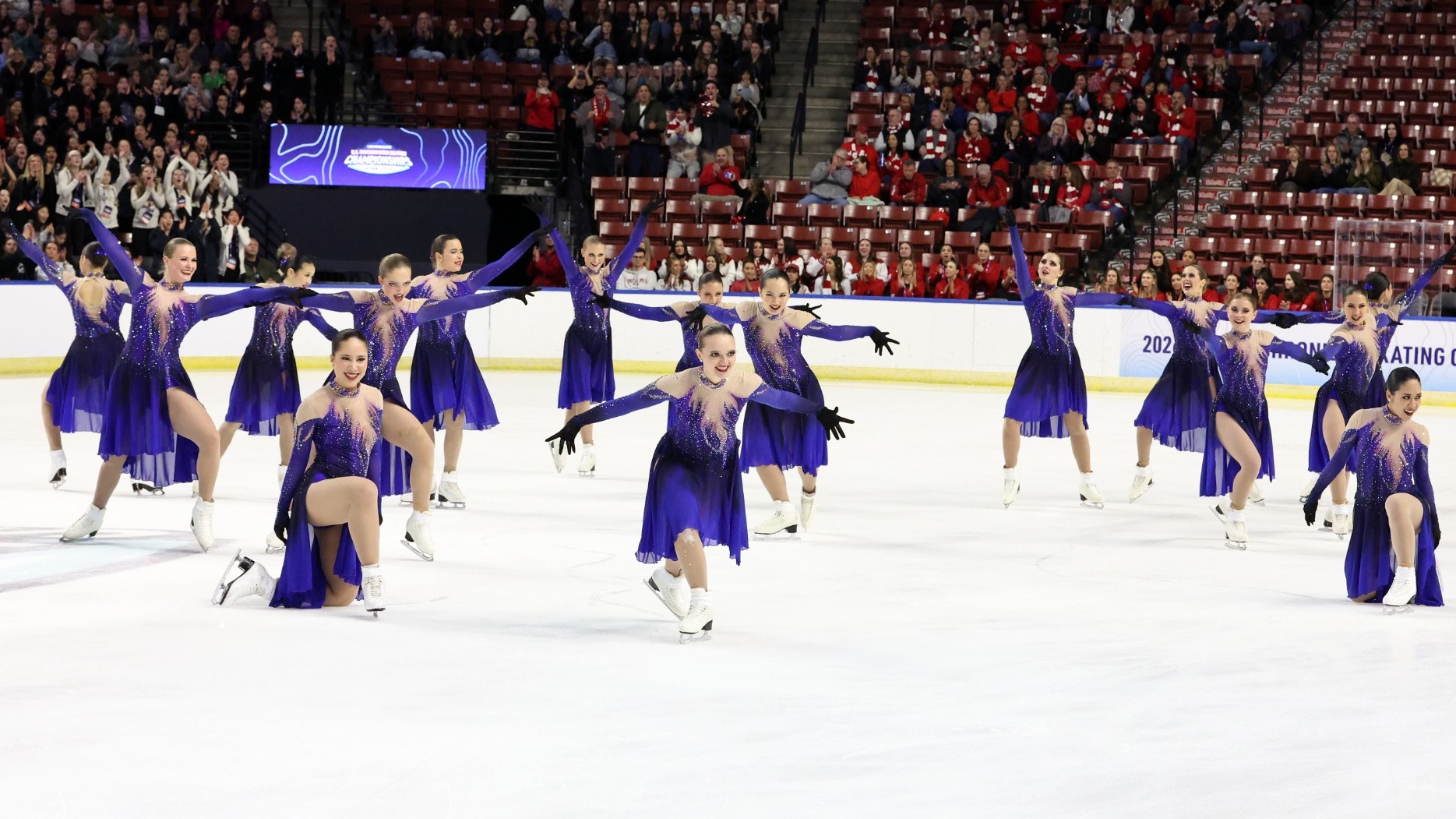 The Haydenettes strike a pose during their short program with their arms extended and looks of determination on their face. The athletes are wearing a long sleeve purple skating costume with purple and pink detailing on the front resembling flames and a long purple and black ombre skirt. 