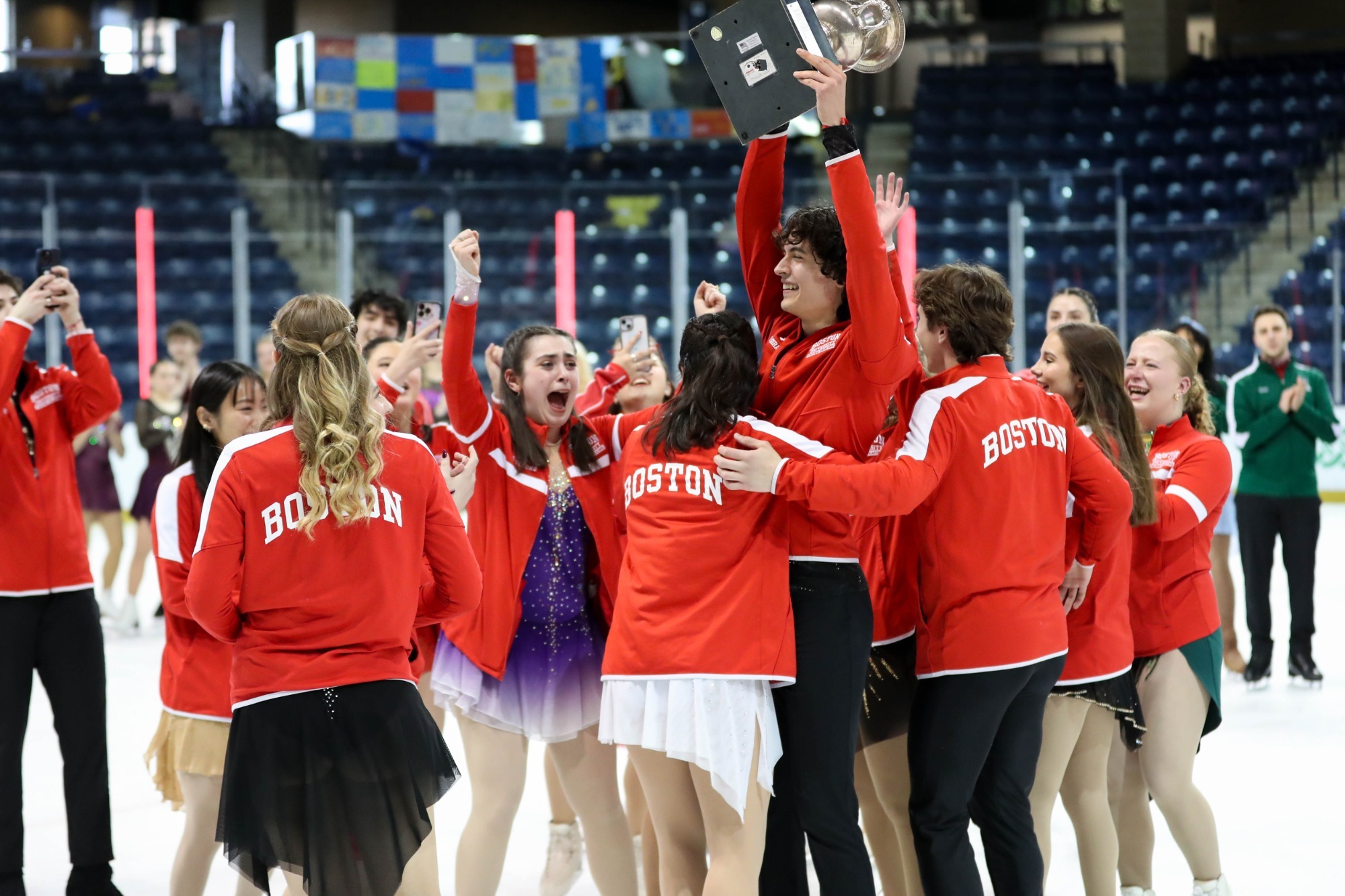 Boston University skaters stand in a circle cheering while one teammate holds a silver trophy up in the air with a smile on his face. The team is wearing matching red jackets with Boston University written in white on the back. 