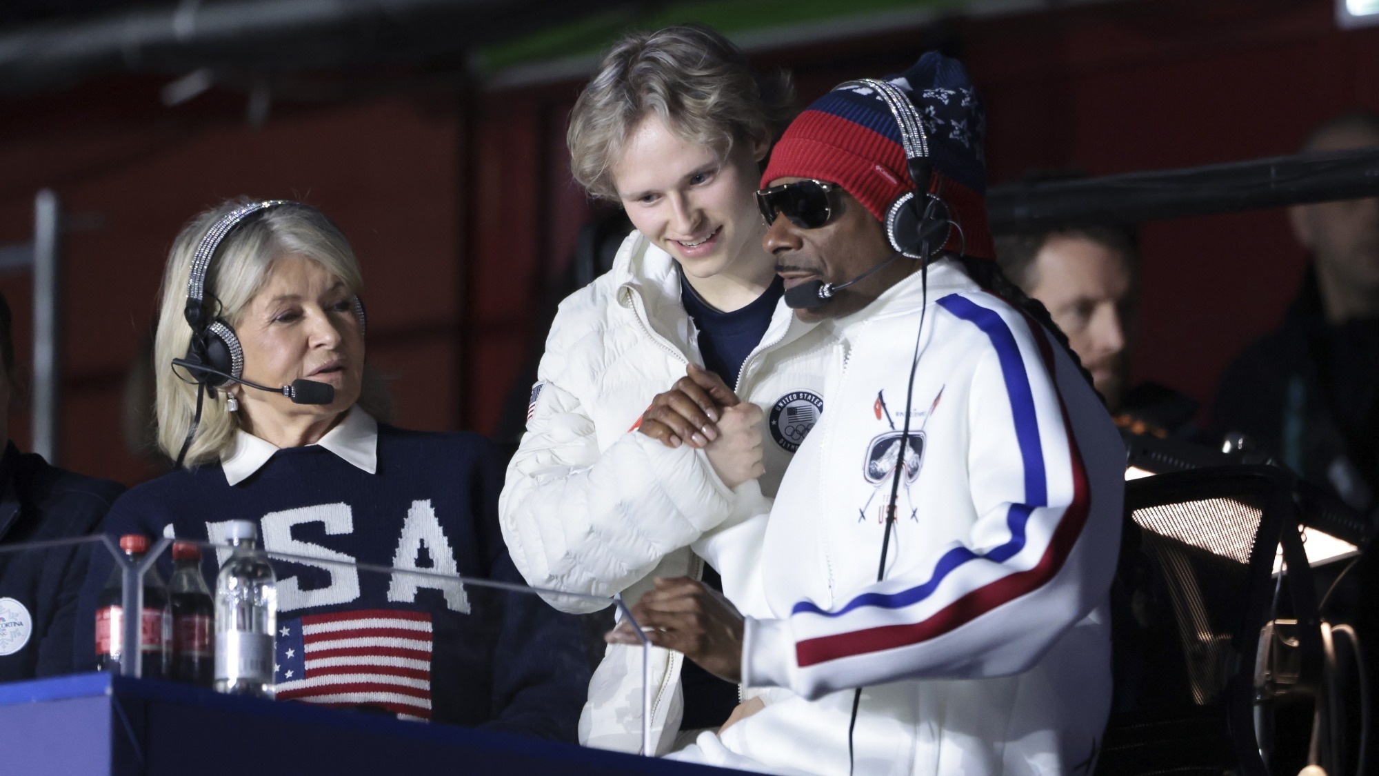 Ilia Malinin, dressed in his white Olympic jacket, stands in the middle of Martha Stewart and Snoop Dogg during the women's final. 