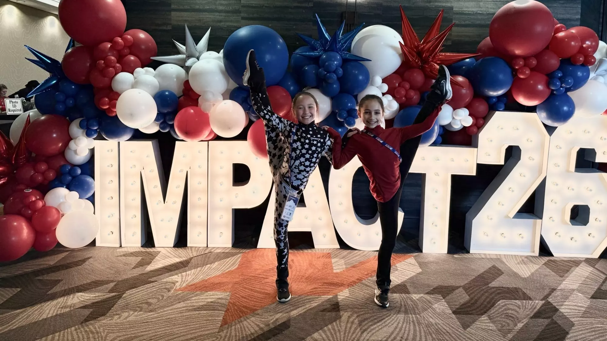 Two young girls pose with one leg up in front of the Impact Summit 2026 sign.