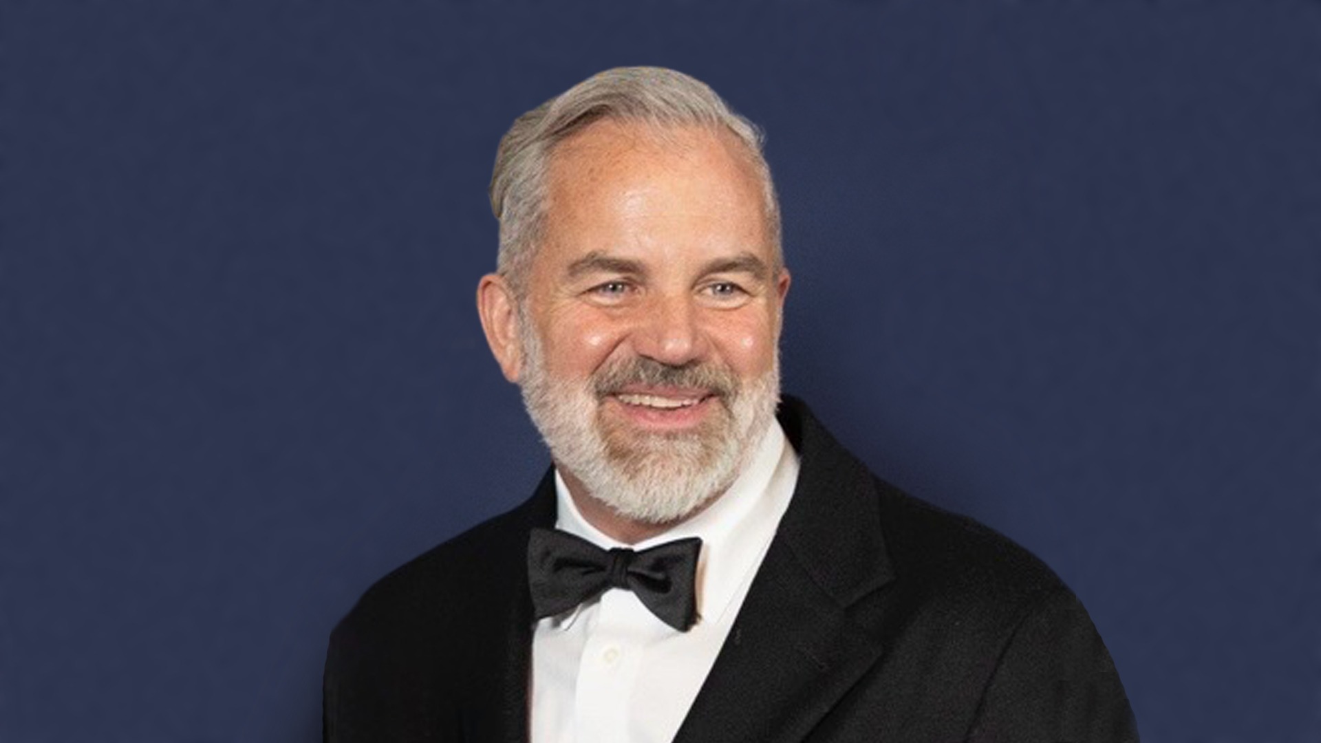 Headshot of Kevin Rosenstein in front of a navy blue background. Kevin is a man with grey hair and a grey beard wearing a black tux with a white dress shirt and black bowtie. 