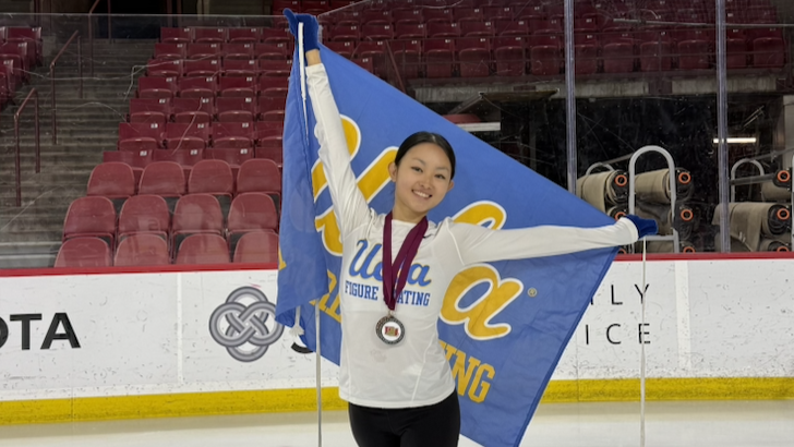 Audrey Lu stands proudly at center ice, wearing a white UCLA Figure Skating long-sleeved shirt, holding a blue UCLA Figure Skating flag and wearing a medal.e UCL