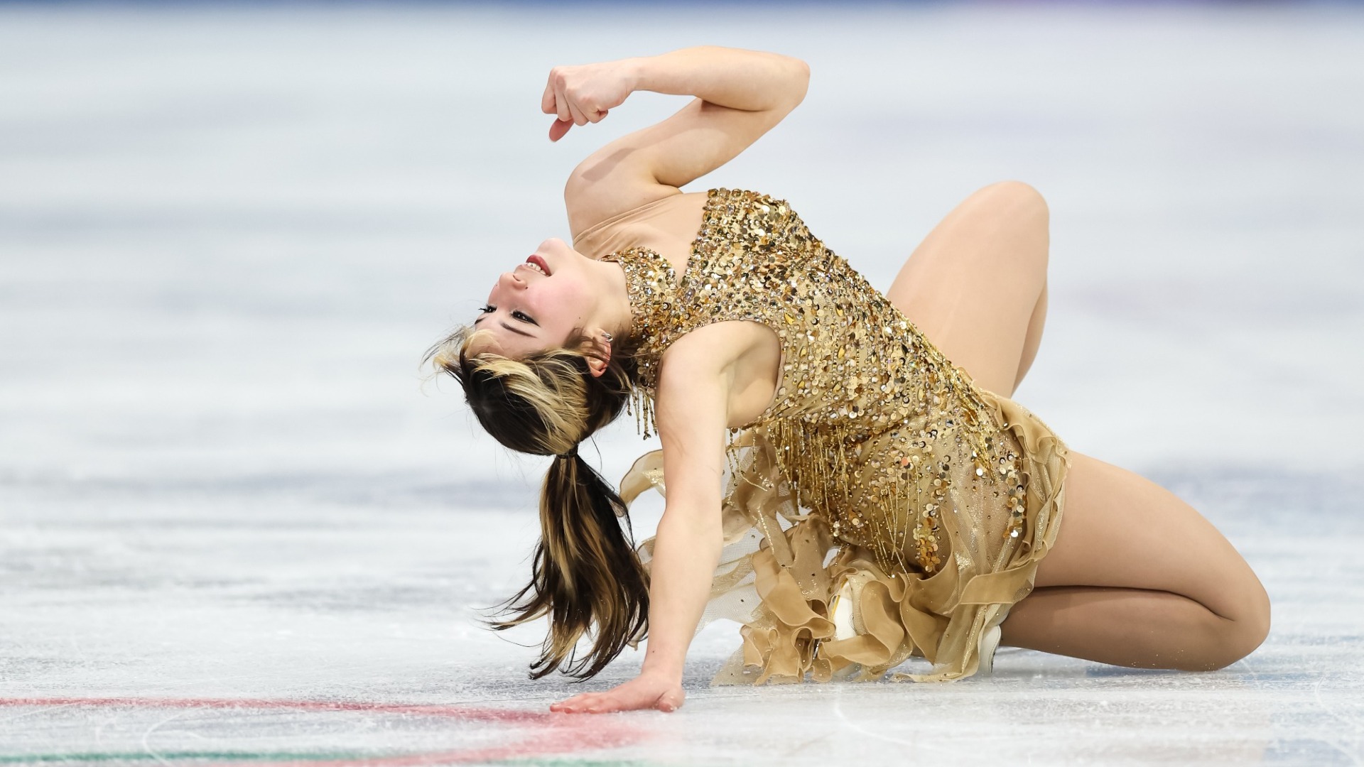 Alysa Liu performs a knee slide with her head leaded back. Alysa is a young woman with brown hair with blonde horizontal stripes in it wearing a one shoulder gold sparkly dress. 