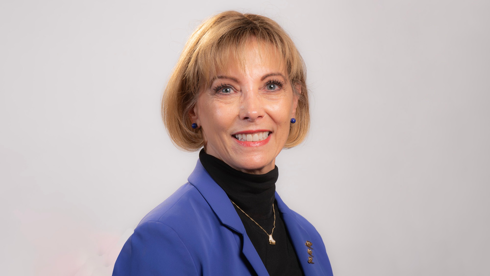Headshot of Heidi DeLio Thibert in front of a white background. Heidi is a woman with shirt blonde hair wearing a black top with a blue blazer over top. She is smiling.