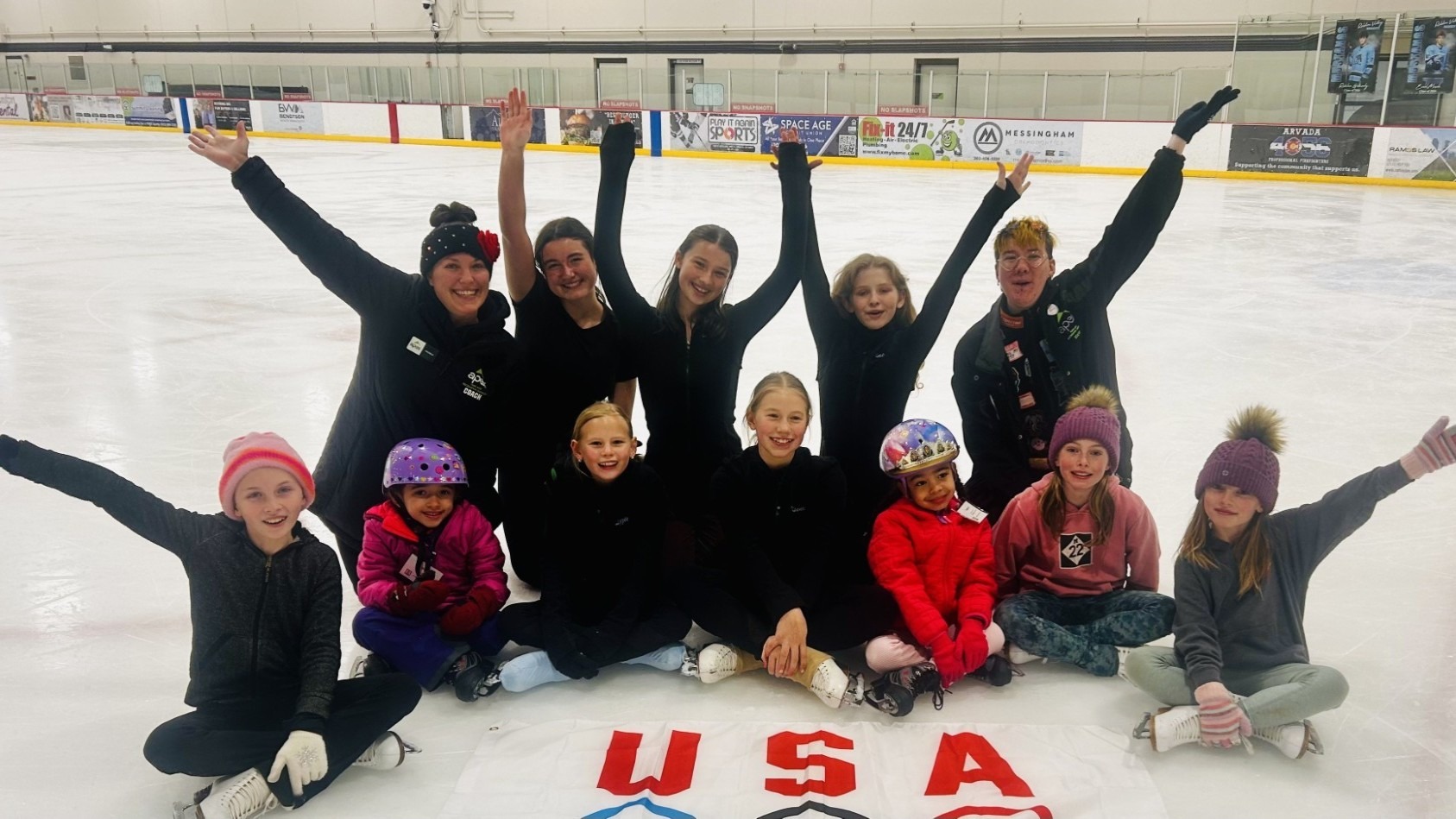 Skaters from Apex Parks and Rec pose with their arms raised and smiles on their faces with a flag bearing the Olympic rings in front of them on the ice. 