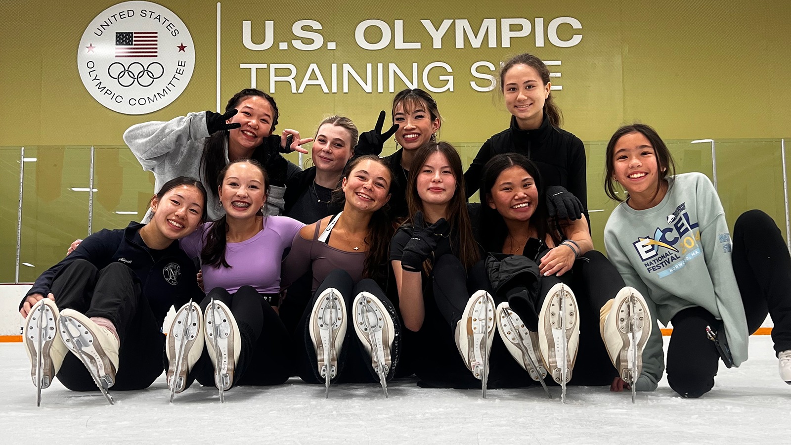 A group of female skaters pose for a photo on the ice on front of a sign that says 