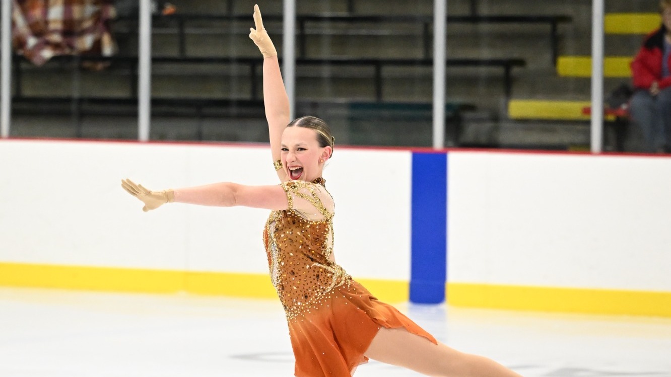 Emmalee Carlson skates with a big smile on her face and both arms raised above her head. She is a young woman with blonde hair tied back in a bun wearing an orange skating costume. 
