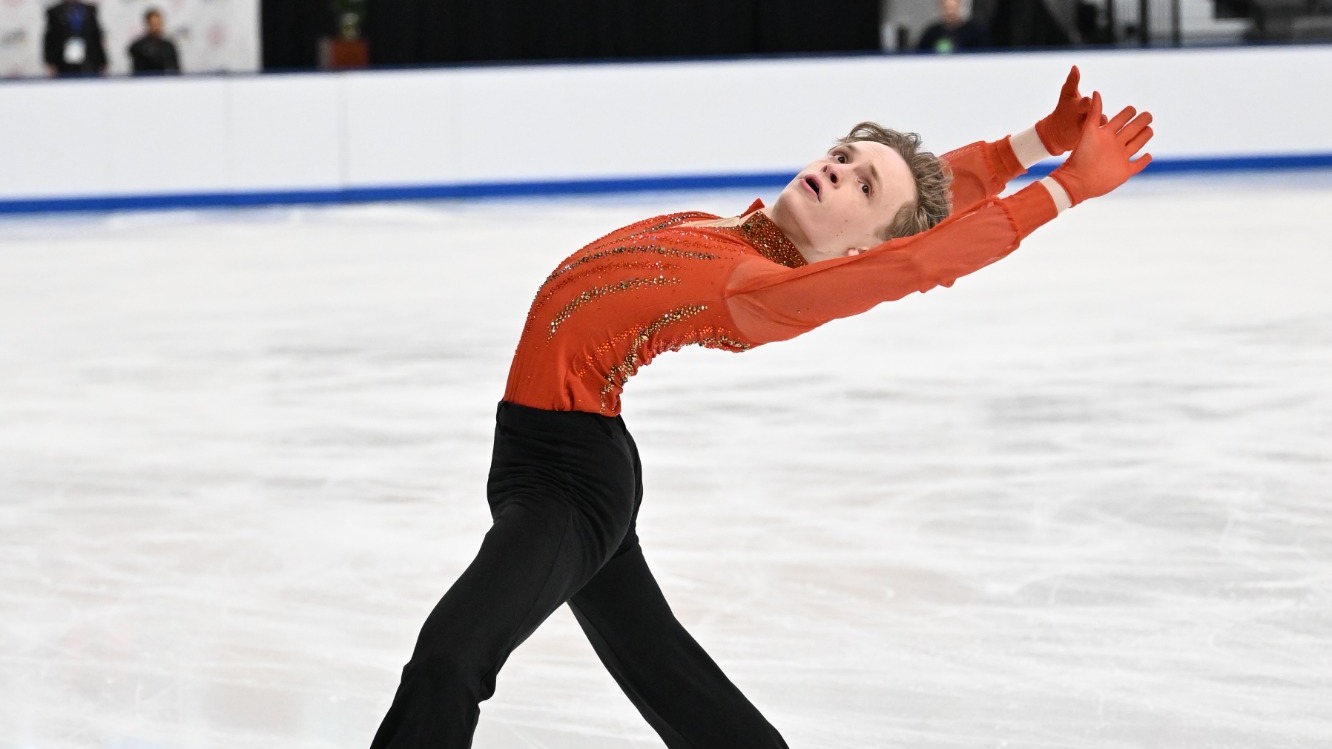 Carson Kandas performs leaning back with his arms extended. Carson is a young man with blonde hair wearing a long sleeve red skating top with jewel detailing down the front and sheer sleeves and black pants. 
