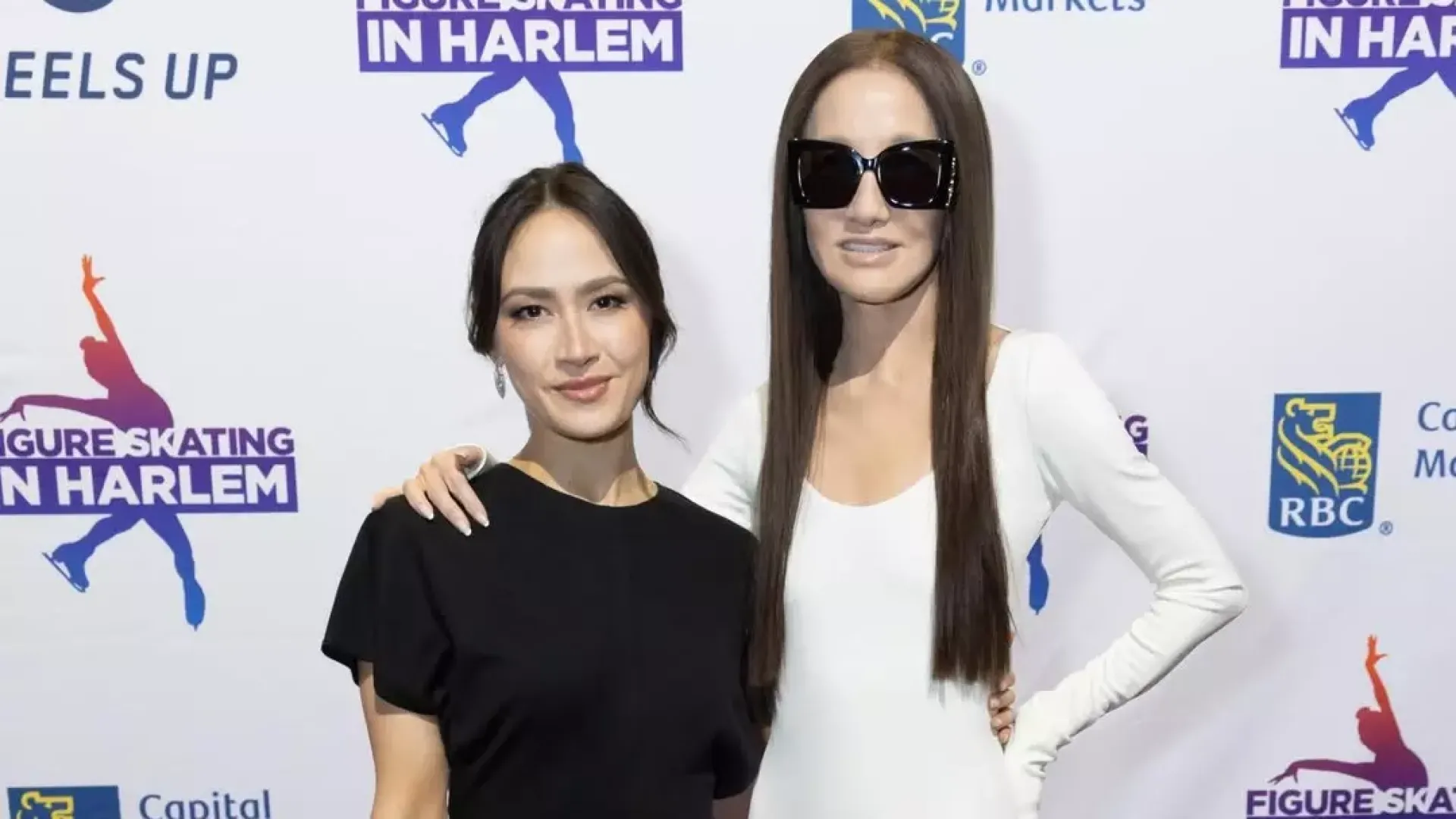 Madison Chock (left) and Vera Wang (right) pose smiling with their arms around each other on the red carpet at the Figure Skating in Harlem gala. Madi is an Asian woman with black hair tied back, wearing long earrings and a floor length short sleeve black dress, black shoes and dangling earrings. Vera is an older Asian woman with long brown hair wearing a long sleeve floor length white dress and black sunglasses. 