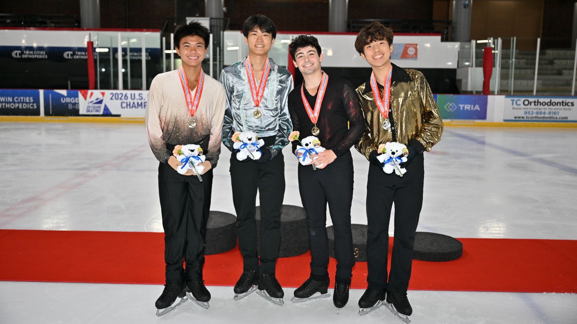 The four U.S. collegiate senior men's medalists stand on the podium with medals around their neck. They are all smiling and holding flowers while wearing their various free skate competition costumes