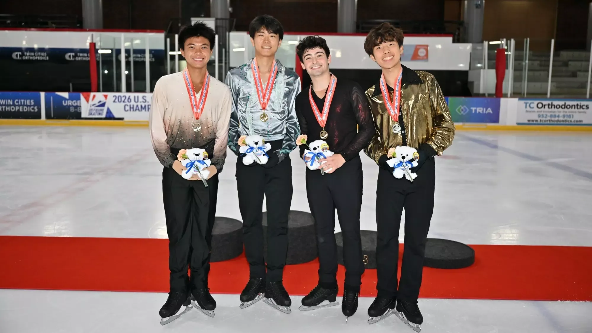 The four U.S. collegiate senior men's medalists stand on the podium with medals around their neck. They are all smiling and holding flowers while wearing their various free skate competition costumes