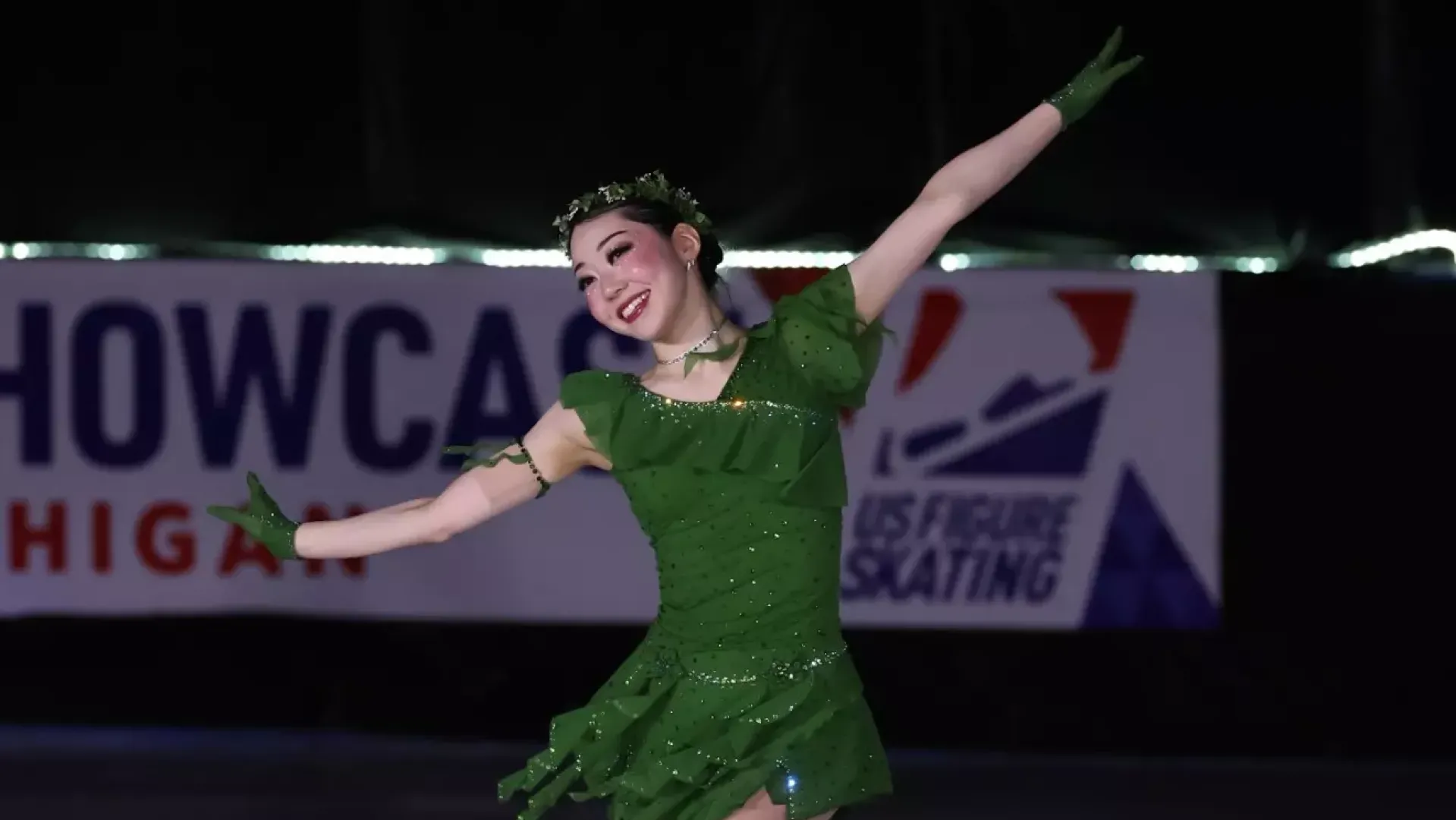 Koko Hirano performs with a big smile on her face and both arms extended outward. She is a young Asian woman wearing a green skating costume with a green leaf headband. 