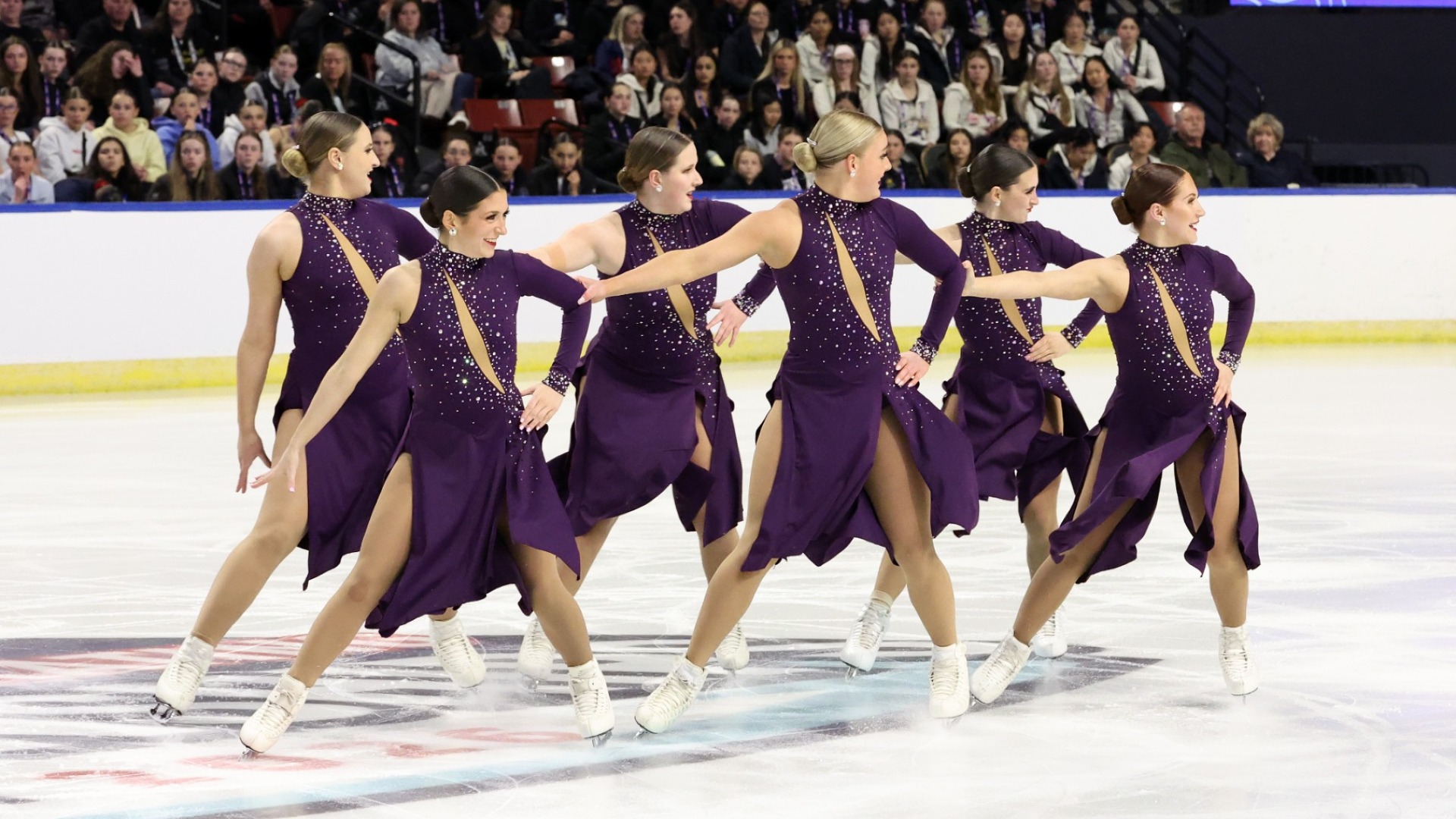 Trine University competes at the 2026 U.S. Synchronized Skating Championships. Six skaters stand at center ice looking to the right in unison. All athletes are wearing coordinating dark purple skating costumes with one long sleeve and crystal detailing down the front. 