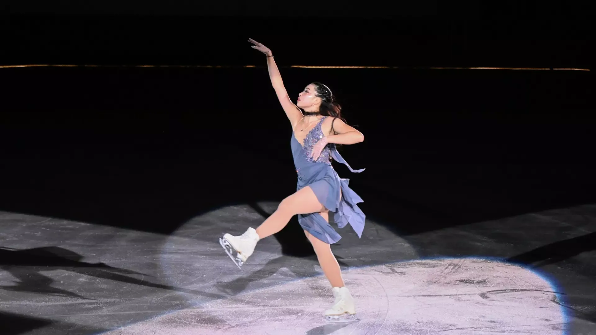Weyna Chen, wearing flowing blue costume, performs a graceful pose on the ice, extending one leg forward and raising one arm overhead under a spotlight.