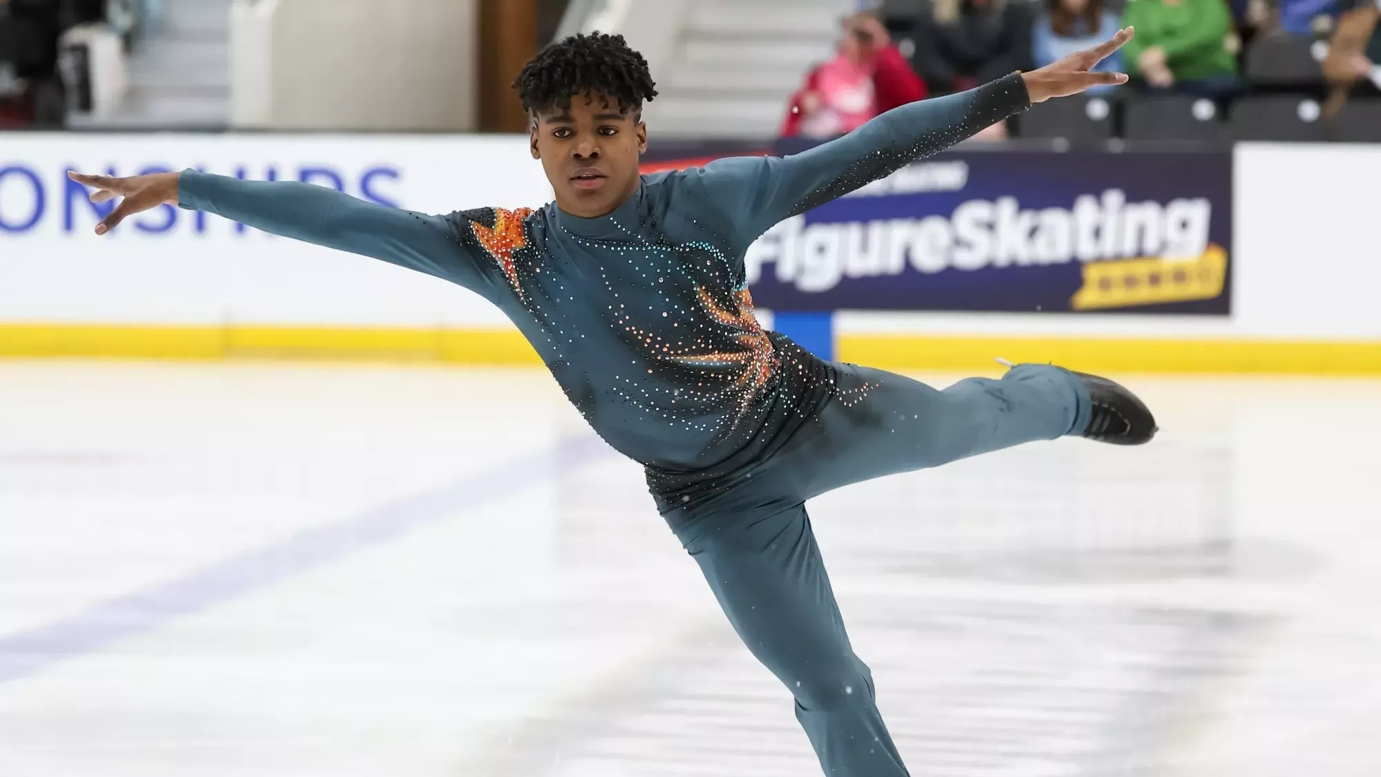 Caleb Farrington, arms extended, skates at the 2026 U.S. Championships. He is wearing a teal long sleeve skating costume with orange detailing on the shoulder and matching teal pants. 