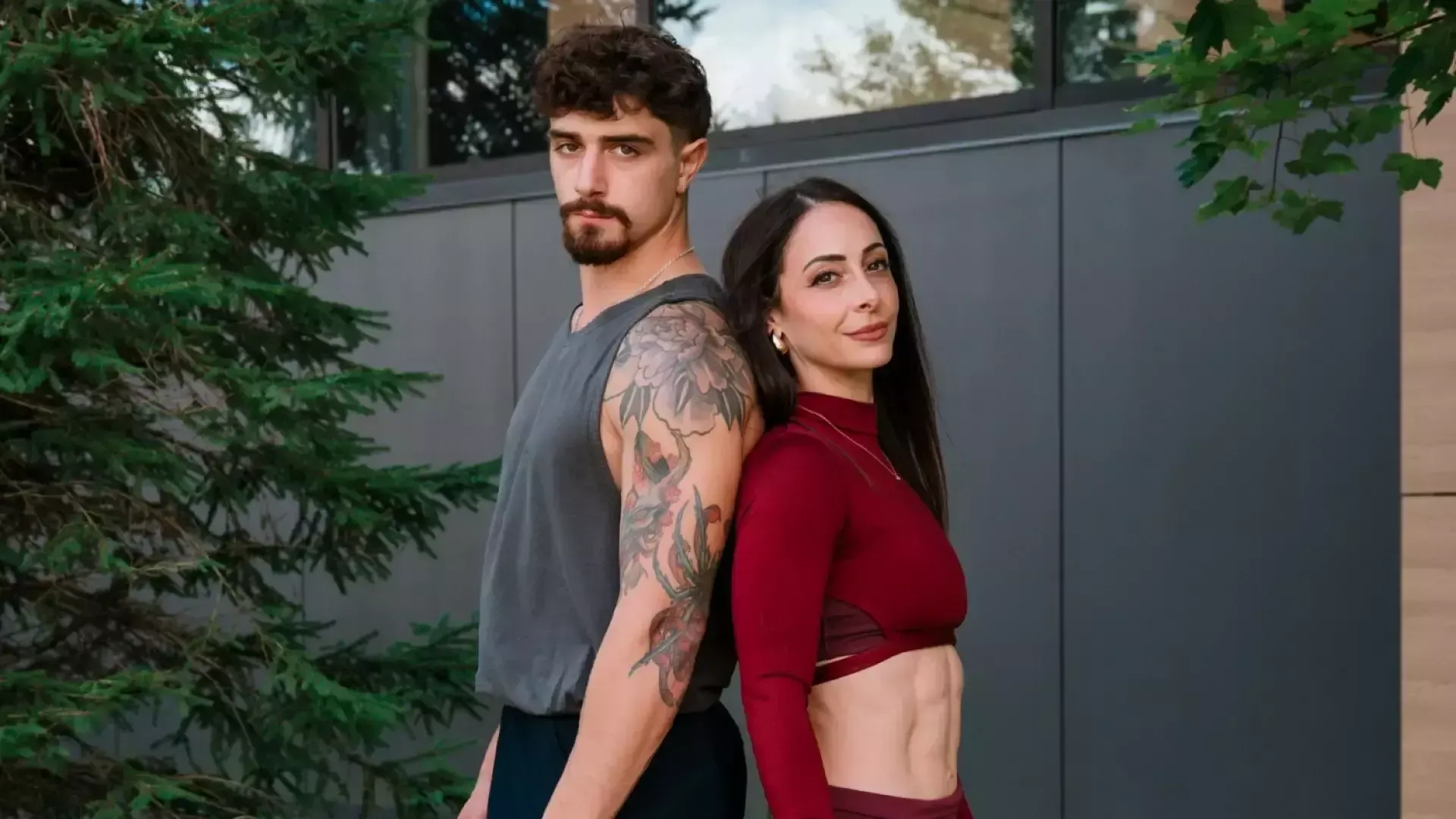 Deanna and Danil stand back to back in front of a grey building, looking directly at the camera. Danil has short, wavy dark hair with a beard and mustache. He wears a gray sleeveless athletic shirt and a thin silver necklace. He has a floral tattoo sleeve. Deanna has long, straight dark-brown hair and looks toward the camera with a subtle smile. She wears a fitted burgundy long-sleeved athletic crop top with a high neckline and matching pants. 