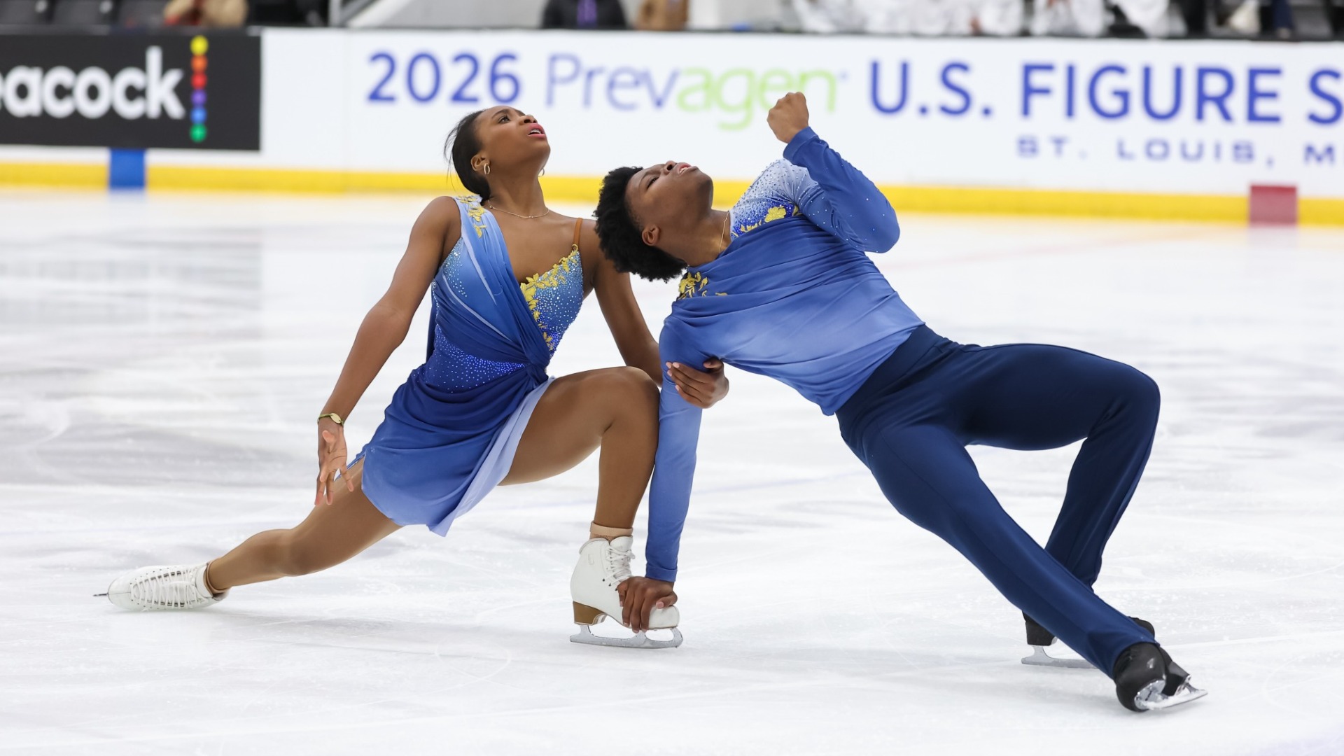 Anaelle Kouevi and Yann Homawoo perform a dramatic synchronized move on an ice rink. Anaelle, wearing a flowing blue dress with yellow and sparkling accents, is in a deep lunge with one leg extended behind her while reaching toward the ice. Beside her, Yann wears a dark blue costume and leans deeply backward, supporting himself on one hand as his other arm bends upward. Both skaters look upward with expressive poses. In the background, spectators sit in the stands