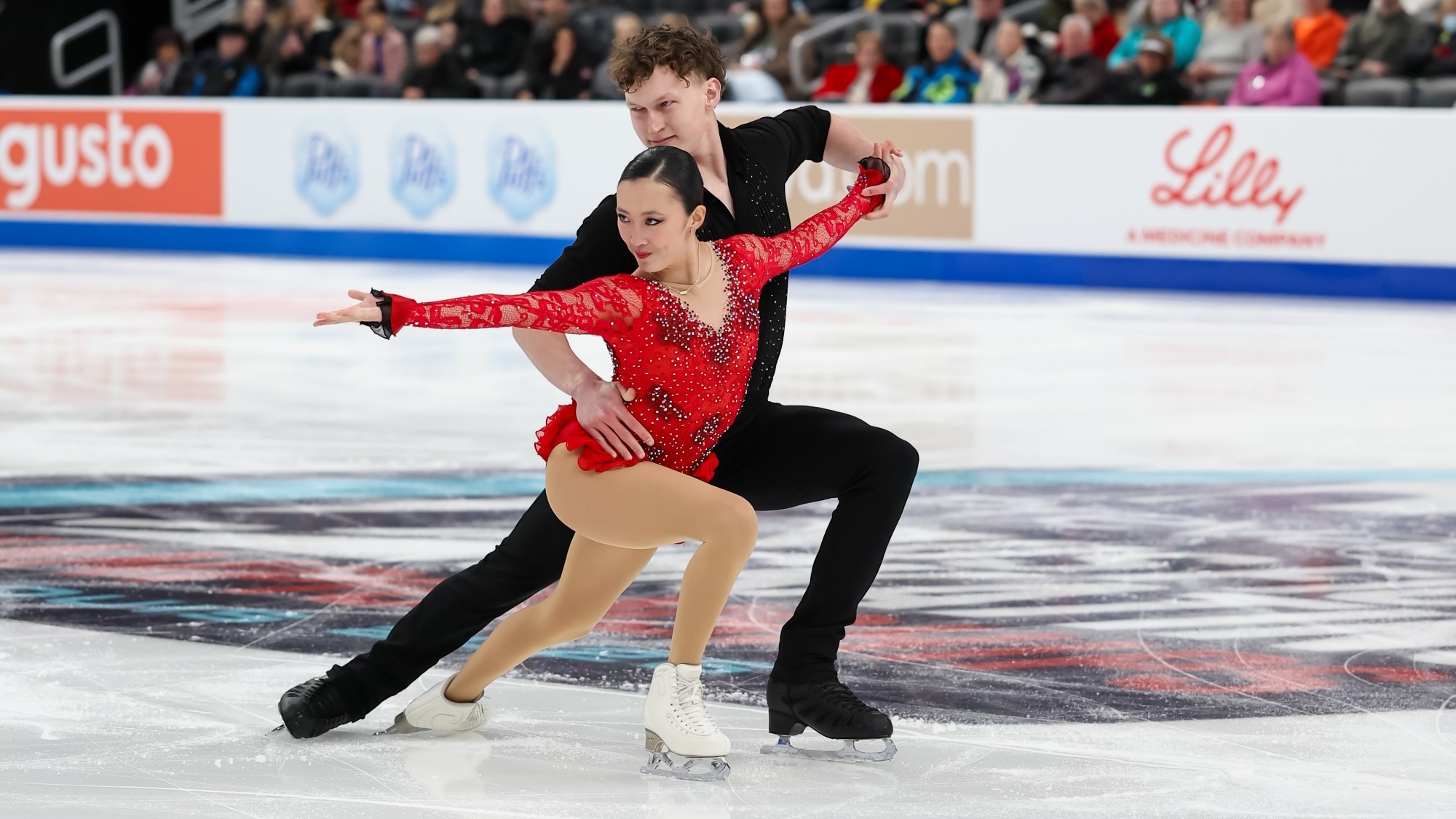 Naomi Williams and Lachlan Lewer perform on the ice. Naomi is in front and has has dark hair pulled back neatly, with a focused, composed expression as she looks toward the left. She wears a sparkling red long-sleeved costume with rhinestone detailing, tan skating tights and white figure skates. Behind her stands Lachlan with short, tousled brown hair. He has a concentrated expression as he looks downward toward his partner, wearing a fitted black skating costume. He supports her around the waist while extending one arm toward her raised hand, and both skaters hold a low, dynamic position as they glide across the ice. Spectators fill the arena seats in the background, with colorful rink advertisements along the boards.