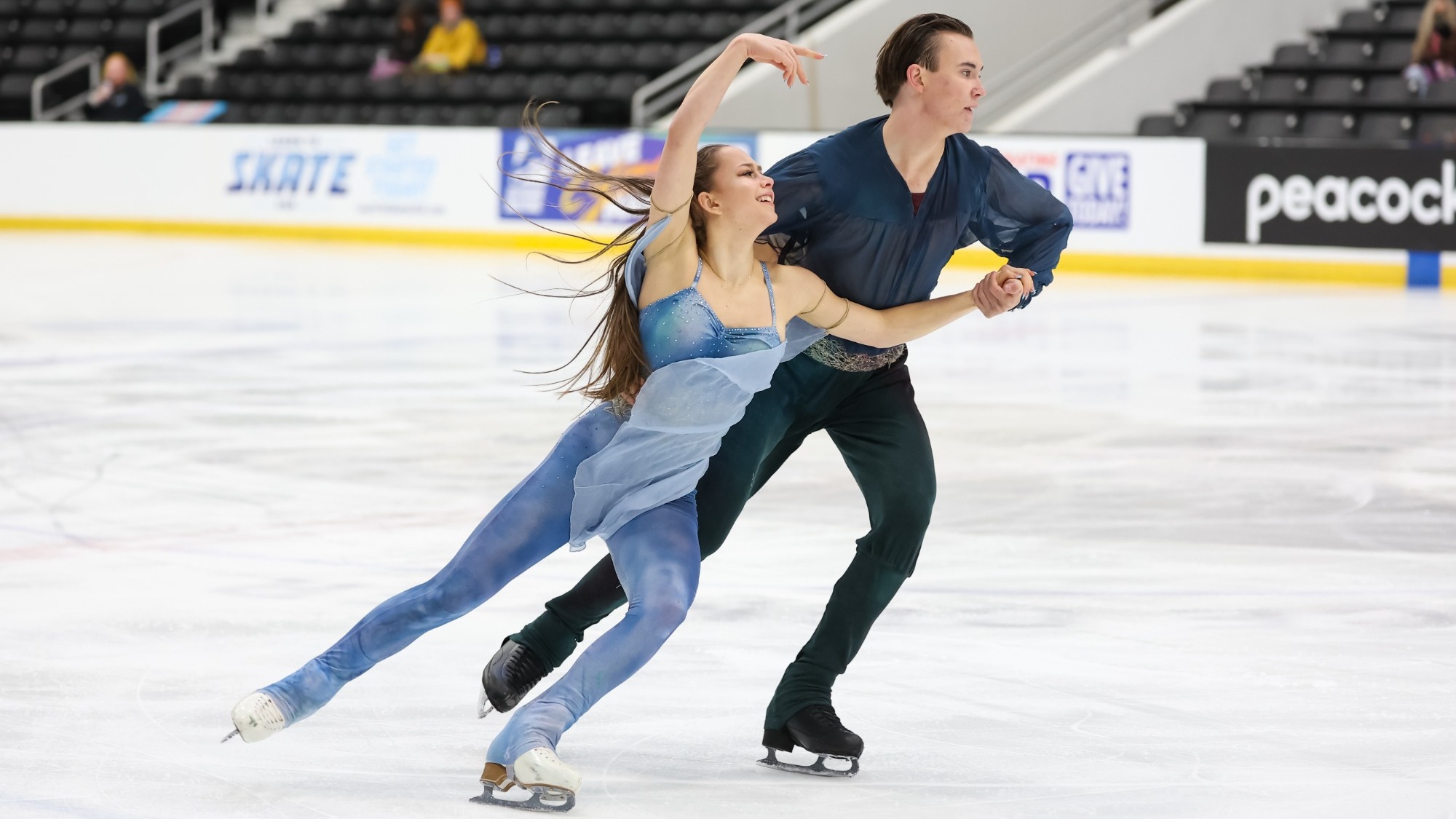 Hana Maria Aboian and Daniil Veselukhin perform together on the ice during a pairs or ice dance routine. Hana Maria wears a light blue, sparkly costume with a flowing sheer skirt and has long, brown hair that streams behind her as she moves across the ice. She raises one arm above her head and extends one leg behind her while looking toward Daniil. Daniil wears a dark blue long-sleeved shirt and black pants, with short, dark brown hair slicked back. He skates alongside her while holding her hand.