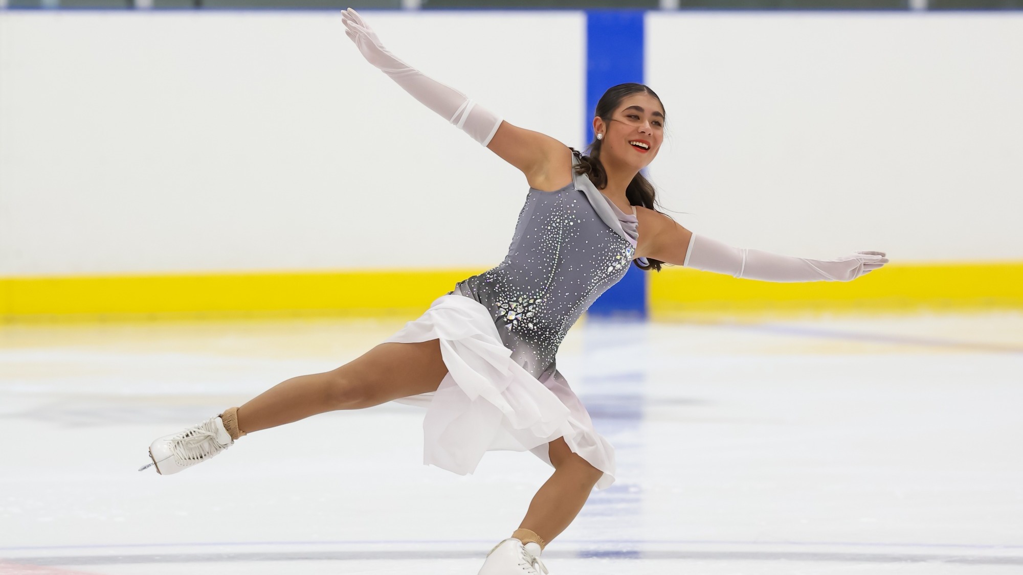 Amal Israilova glides across the ice with one leg extended behind her and both arms outstretched. She wears a gray rhinestone-studded costume with a flowing white skirt and long white gloves, with her dark hair pulled back. She smiles as she performs inside an ice rink.