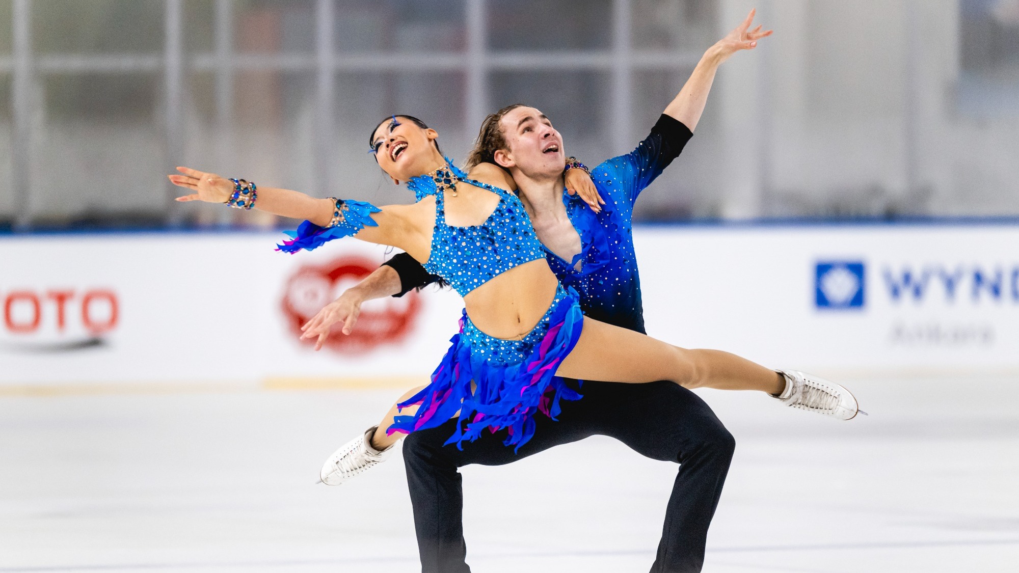 Effie Chen and Gordei Chitipakhovian perform a dramatic synchronized move on the ice. Effie in front wears a bright blue, rhinestone-covered costume with blue and purple fringe, while Gordei behind her wears a dark blue, rhinestone-covered long-sleeved shirt and black pants. Effie extends one leg horizontally to the side and stretches both arms outward, while Gordei supports her in a low, wide-legged position with one arm raised overhead. Both skaters look upward with expressive faces. The ice rink and advertising boards are visible in the background.