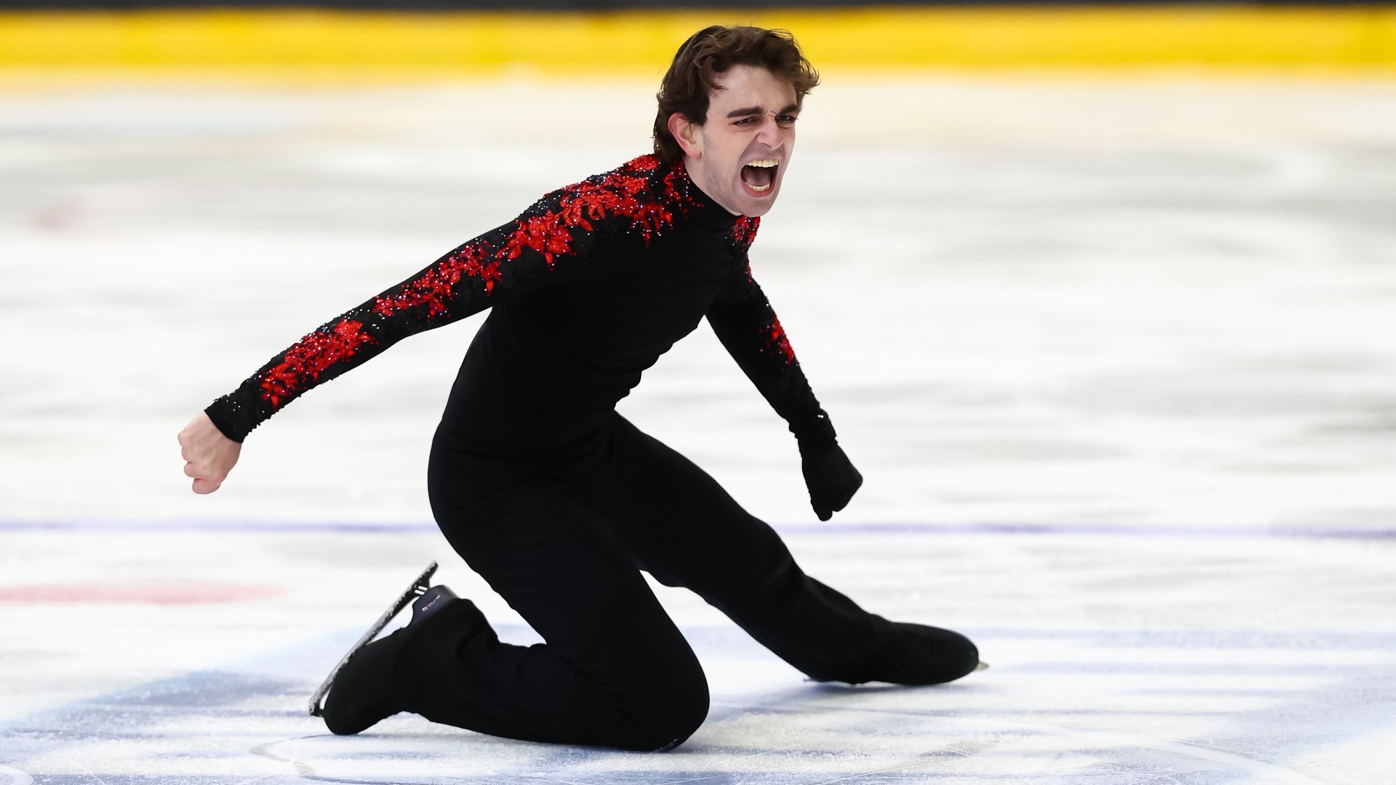 Lucas Appel kneels on the ice with his arms outstretched with a look of determination on his face. He is a man with short, curly brown hair wearing a long sleeve black top with red detailing down the sleeves, black pants and black skates