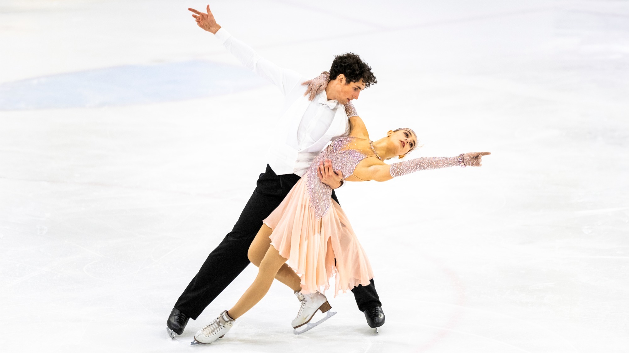 Aneta Vaclavikova and William Lissauer partnered move on a white ice rink. William, wearing a white long-sleeved shirt and black pants, leans over while supporting his partner around the waist and upper back. His left arm is extended upward and outward. Aneta wears a sparkling, pale pink, sleeveless dress with a flowing skirt and matching glittering details. She leans back, extending her right arm outward and pointing with her hand. 