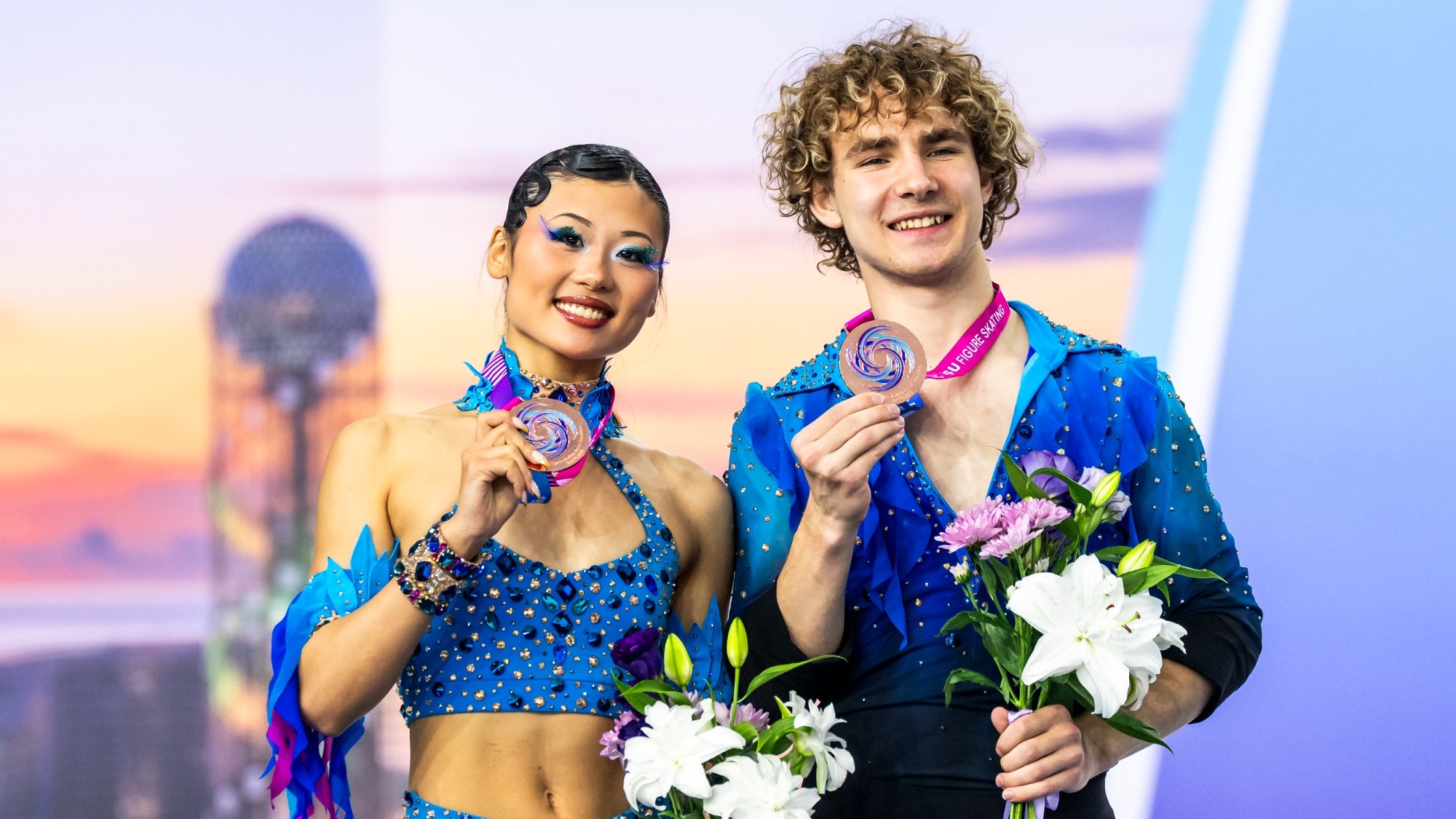 Effie Chen and Gordei Chitipakhovian stand side by side on a brightly lit awards podium, smiling and holding bronze medals and bouquets of white and purple flowers. Effie (left) wears a sparkling royal-blue competition costume with rhinestone embellishments, a cropped halter-style top, matching skirt, and blue-and-purple ribbons trailing from her outfit. She has dark hair tied back in a bun. Gordei (right) has curly light-brown hair and wears a coordinating dark-blue, rhinestone-covered skating costume. He holds his medal up with one hand and a bouquet in the other. Behind them is a large colorful event backdrop. 