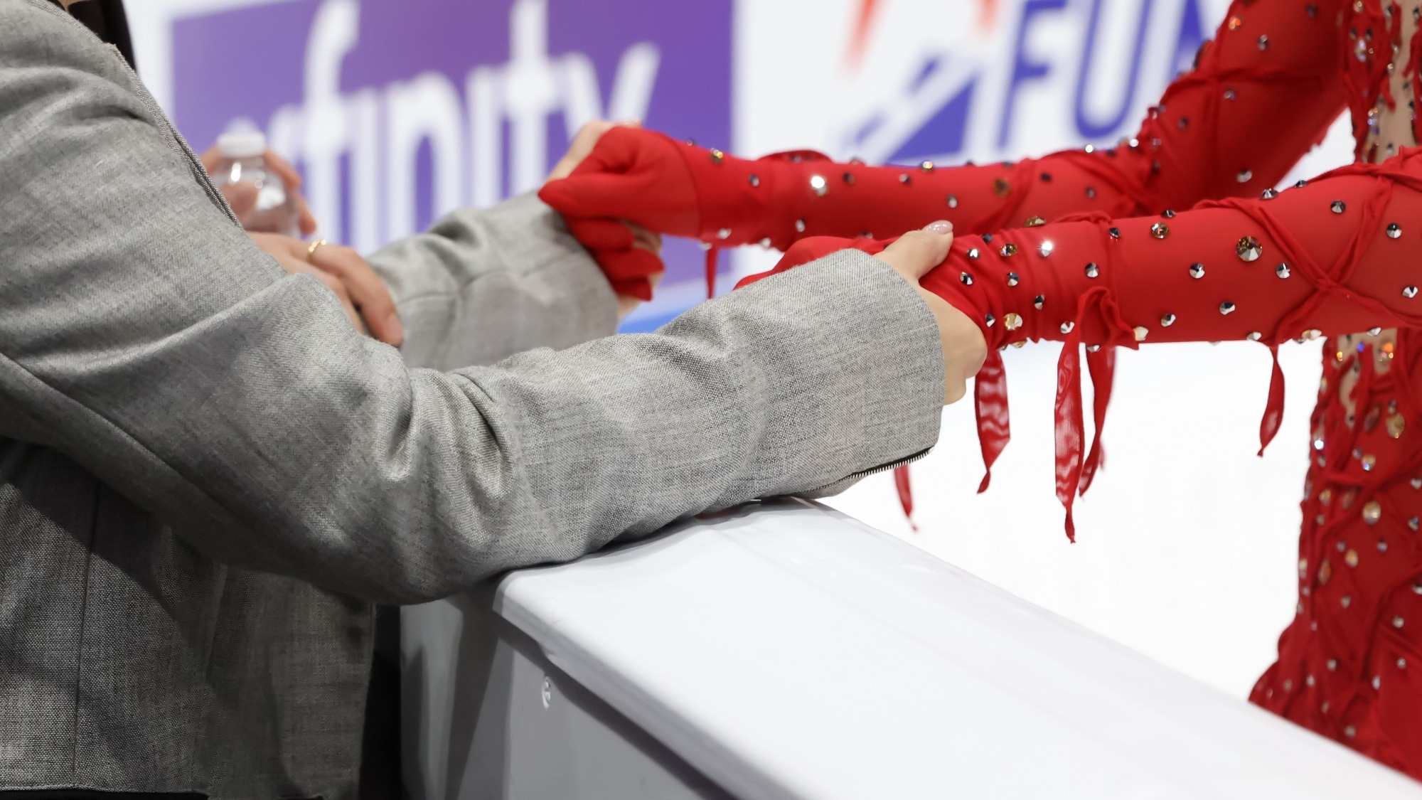Close up photo of a coach wearing a light grey suit jacket holding hands with their skater in support. The skater is wearing a long sleeve red skating costume with matching gloves