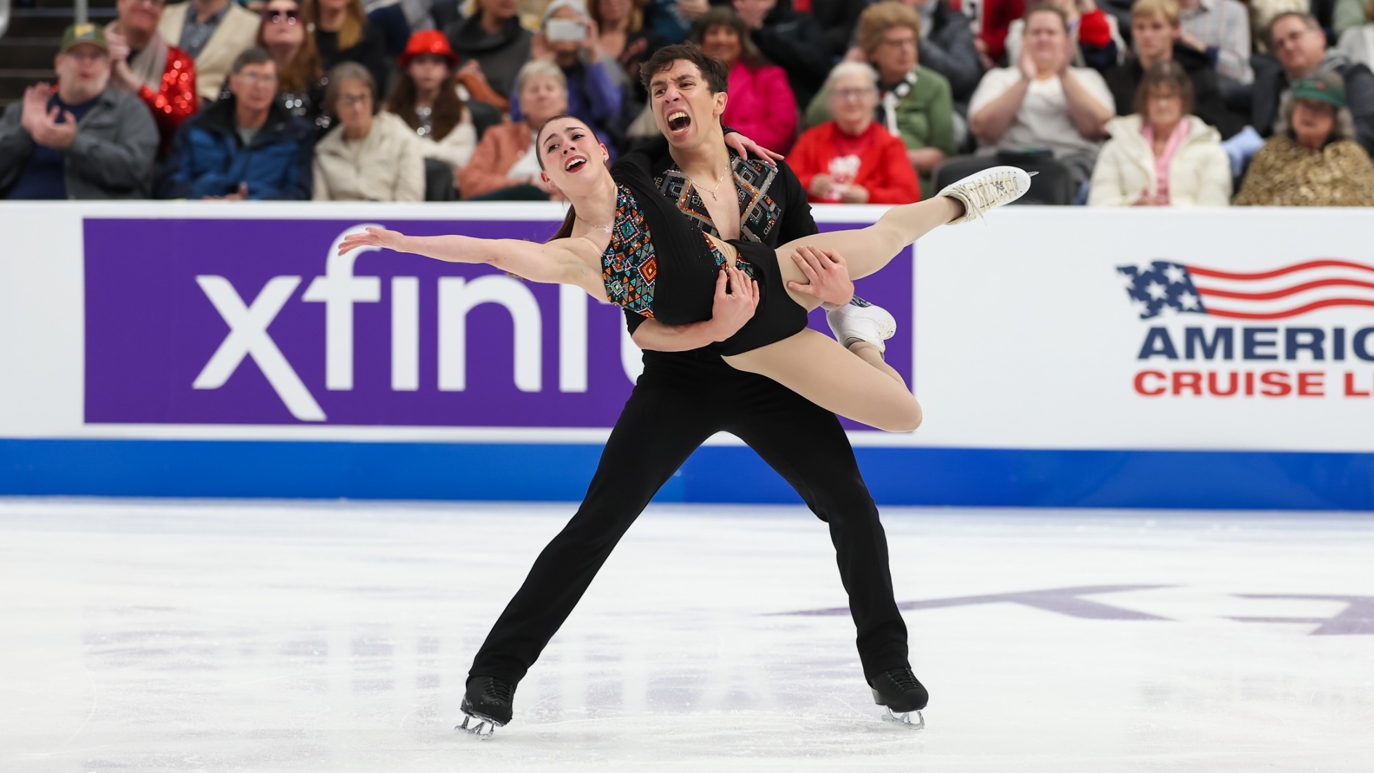 Valentina Plazas and Maximiliano Fernandez perform a pairs routine at the U.S. Championships. Max supports and lifts Valentina. She has one leg extended and one arm outstretched. Both wear black costumes with colorful geometric detailing. Spectators fill the stands in the background.