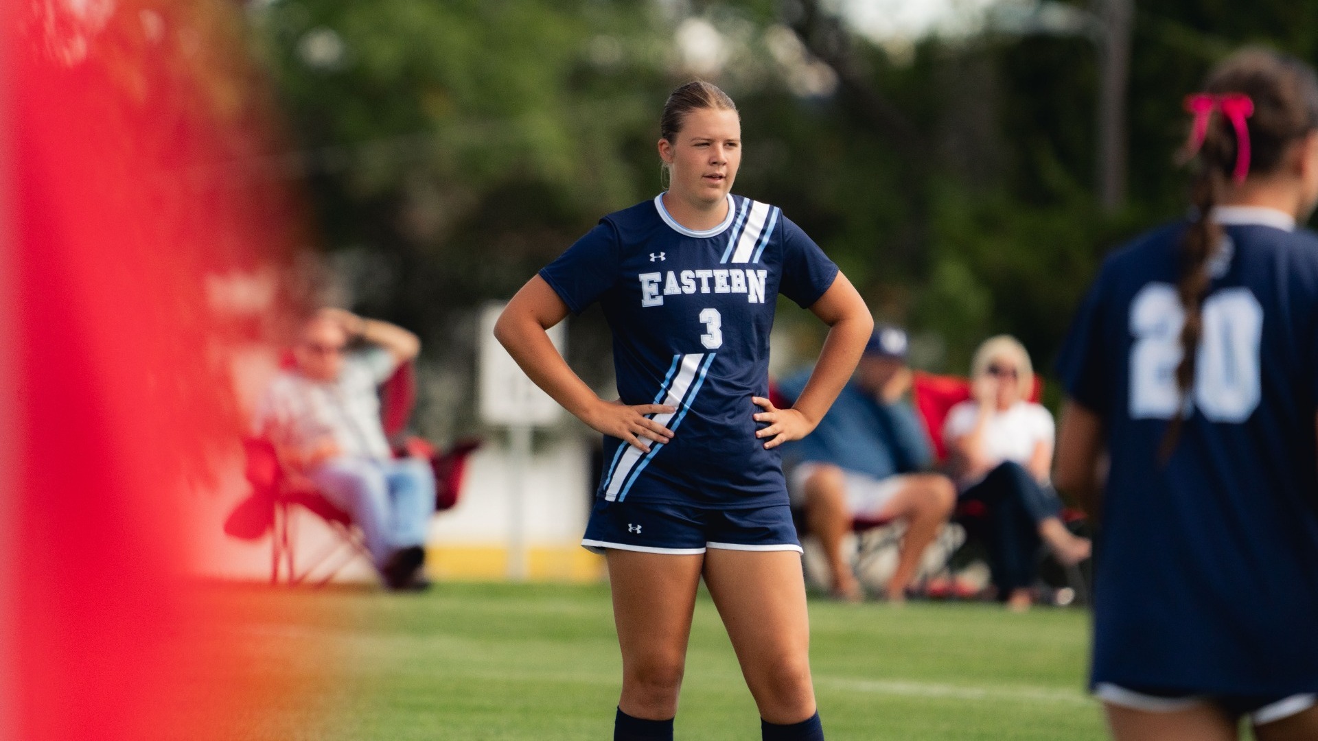 An outdoor action photograph capturing a Utah State Eastern women's soccer player in a dark navy jersey (number 3) standing with her hands on her hips on the grass pitch. A teammate (number 20) is visible in the right foreground, while spectators relax in lawn chairs along the sideline in the blurred background.