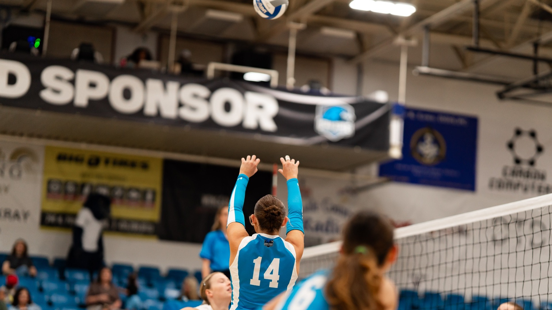 An indoor action photograph showing a Utah State Eastern volleyball player (number 14) in a blue and white uniform leaping near the net with extended arms to set the volleyball. Teammates are positioned around her on the court, with blue arena seating and sponsor banners lining the gymnasium in the background.