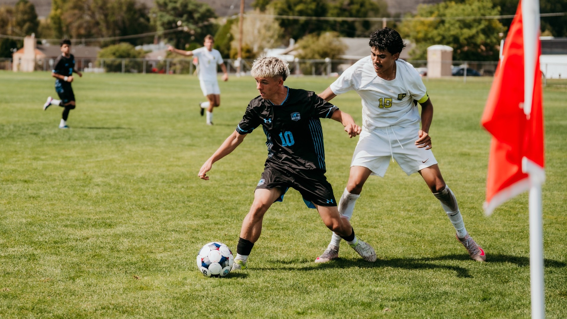 An outdoor action photograph of a Utah State Eastern men's soccer player with frosted-tipped hair (number 10) in a black kit dribbling the ball past an opposing player in white (number 15) near a red corner flag. A green grass pitch stretches across the foreground, with trees and a hillside landscape under a bright sky in the background.
