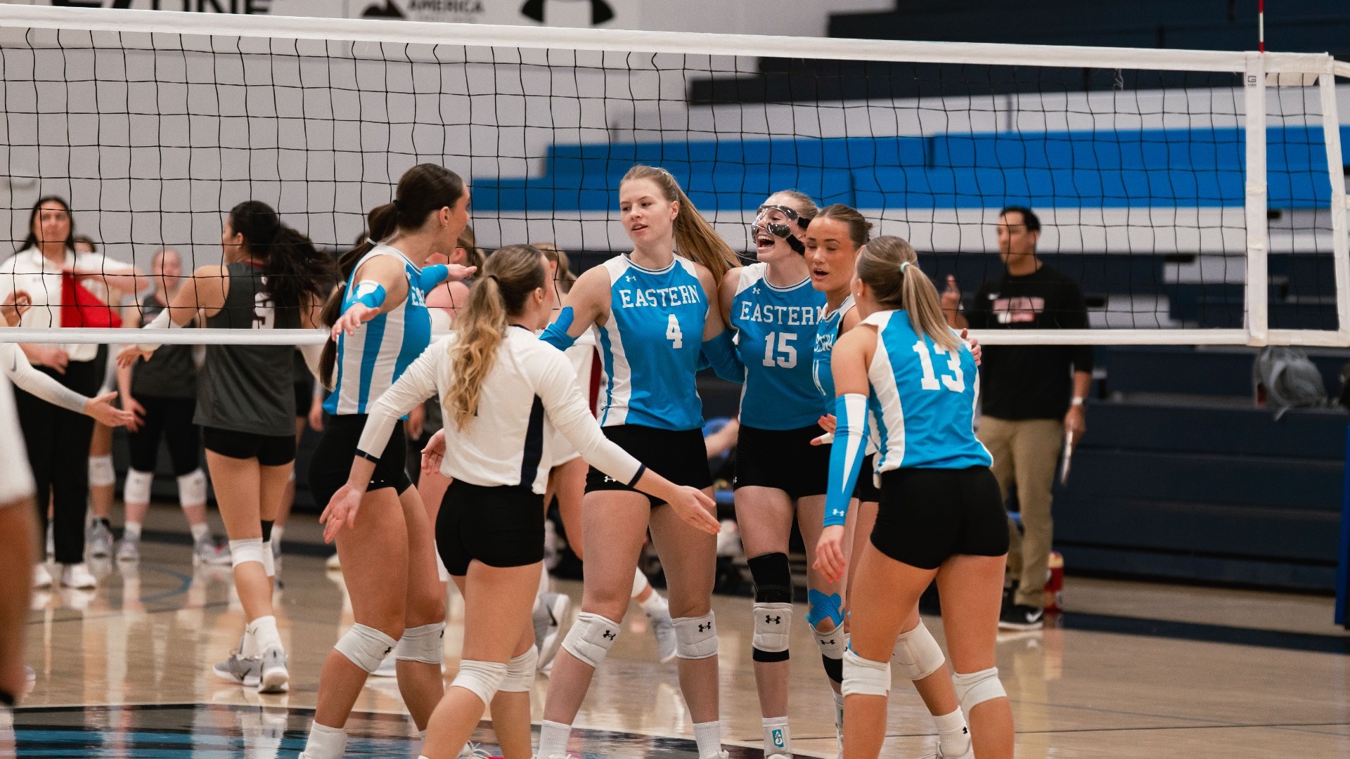 An indoor action photograph capturing Utah State Eastern volleyball players celebrating together on the court. Players in light blue and navy uniforms (including numbers 4, 15, and 13) gather in a huddle, smiling and congratulating one another, with the net, gym flooring, and blue bleachers visible in the background.