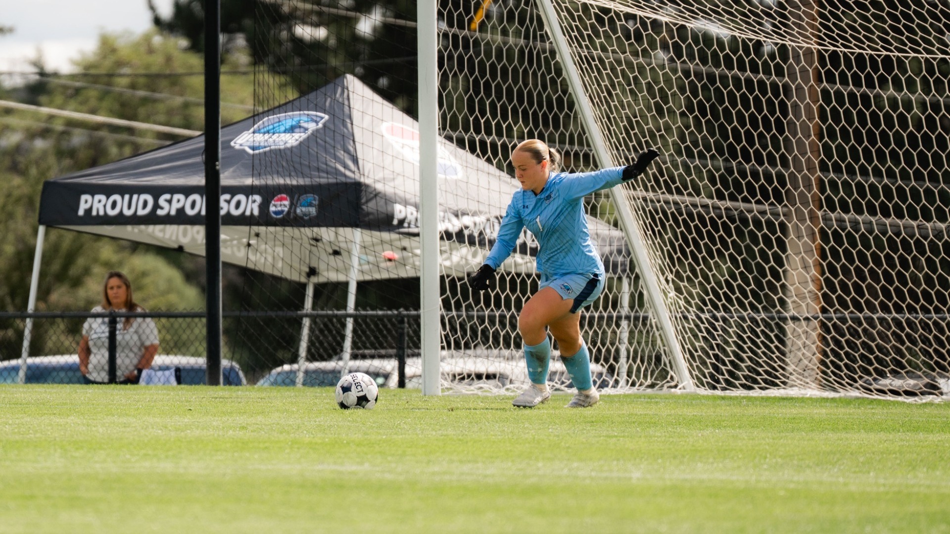 An outdoor action photograph of a Utah State Eastern women's soccer goalkeeper in a light blue jersey set in a athletic stance inside the net frame, tracking a soccer ball resting on the grass. A black Utah State Eastern team pop-up canopy with 