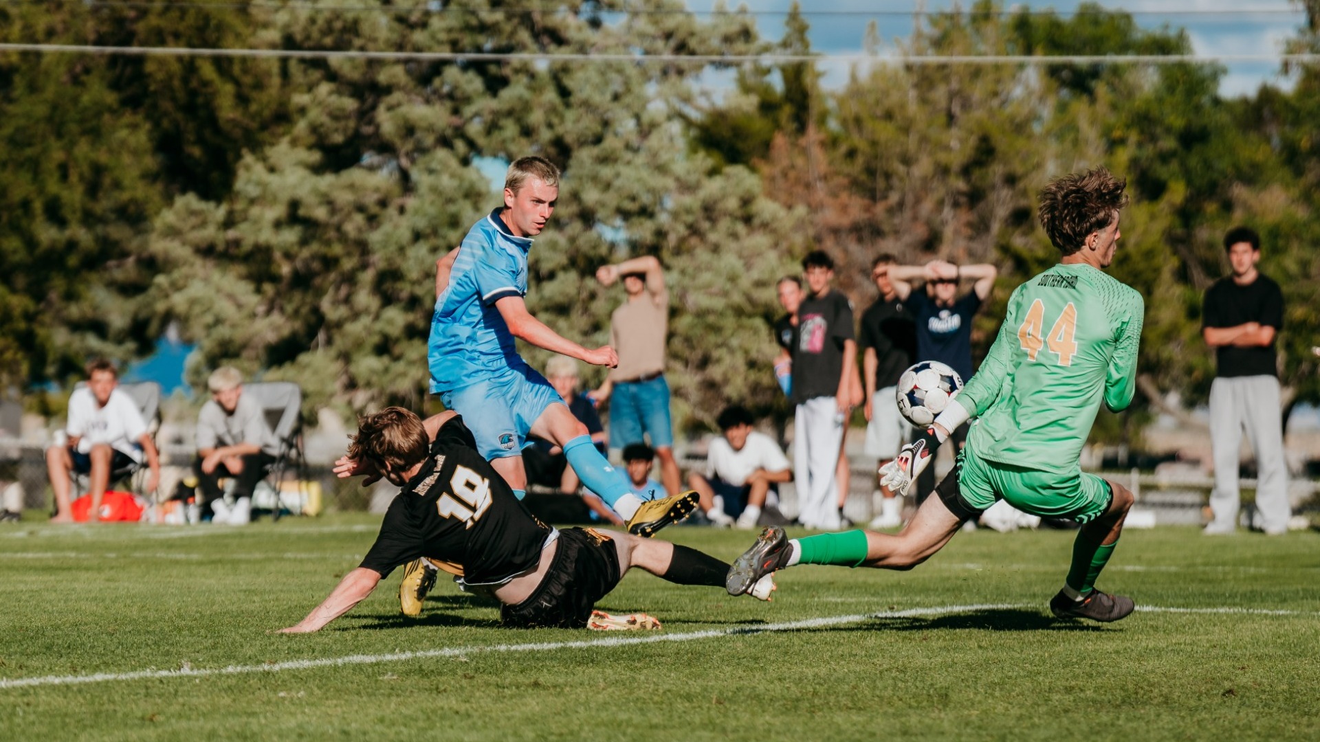 An outdoor action photograph capturing a Utah State Eastern men's soccer player in a light blue kit striking the ball toward the net as an opposing player in black slides across the grass to block the attempt. The opposing goalkeeper in a bright green kit (number 44) lunges toward the airborne soccer ball near the goal line, with spectators lining the background field.