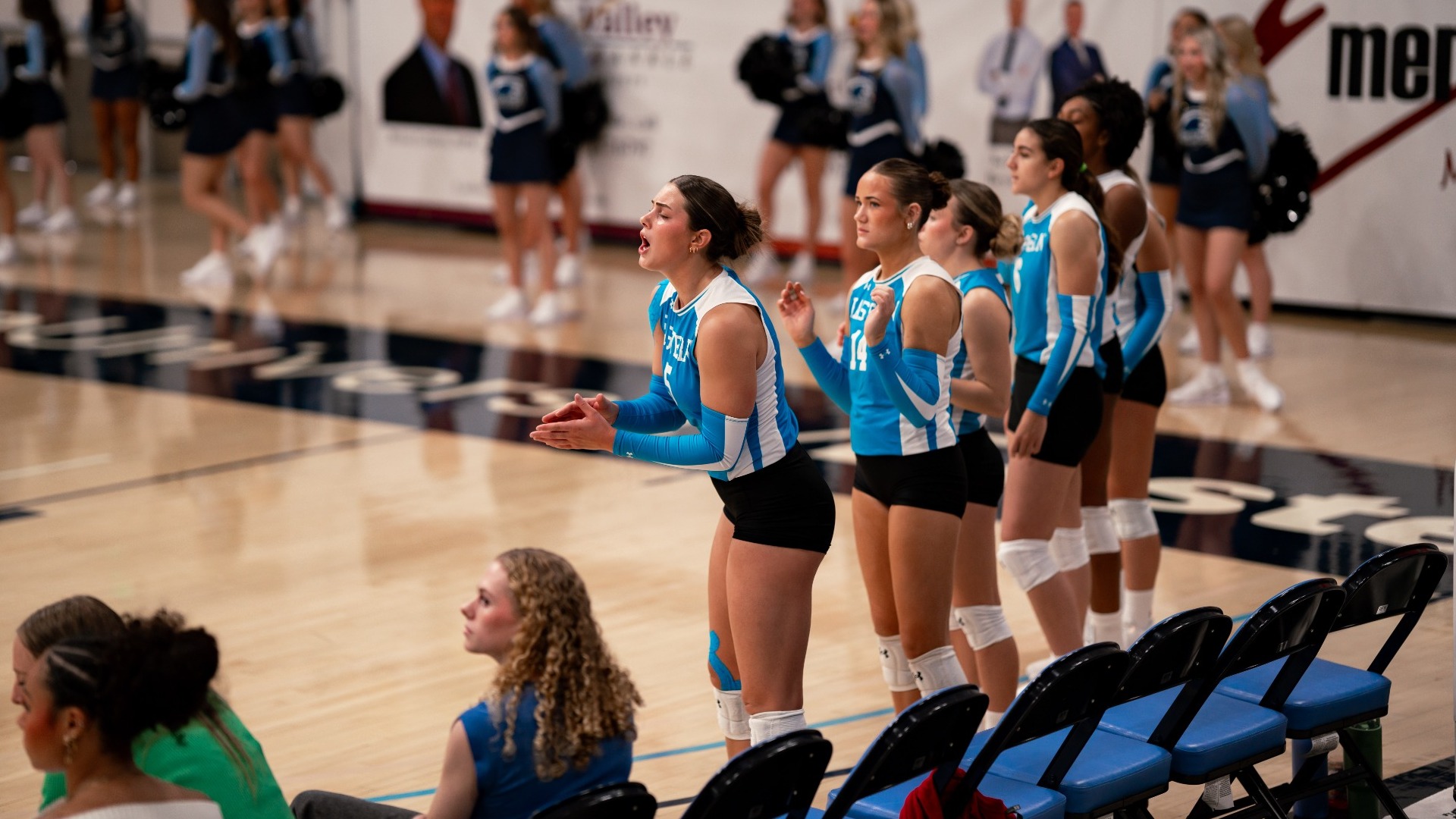 An indoor sports photograph showing a line of Utah State Eastern women's volleyball players standing along the court boundary in their light blue and white jerseys, reacting with focused expressions to the action on court. Cheerleaders and audience members are visible along the gym floor in the background.