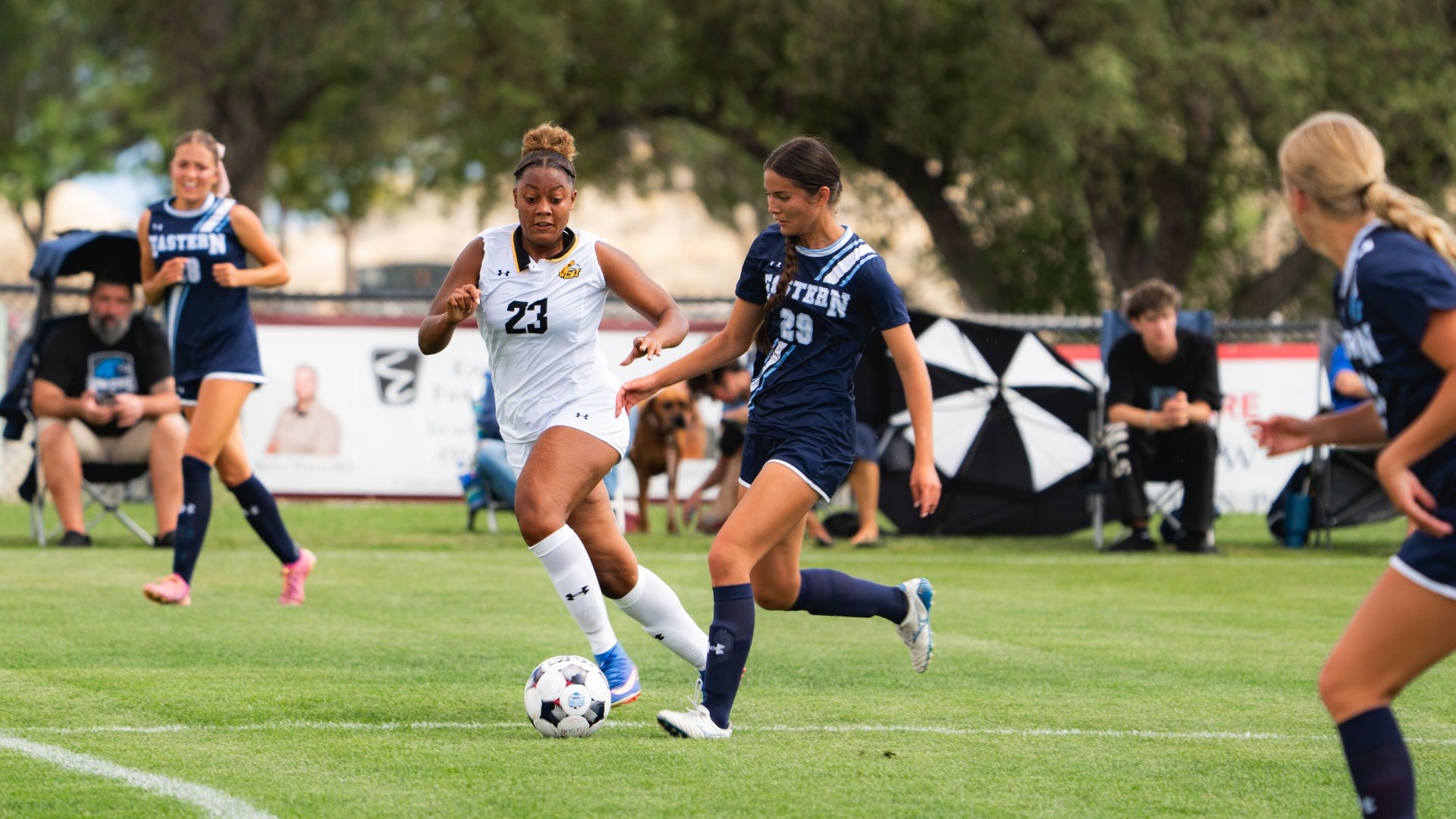 An outdoor action photograph capturing Utah State Eastern defender (number 29) in a dark navy kit pressing closely to tackle the ball away from a College of Southern Idaho opponent wearing a white jersey (number 23). Teammates in navy kits track the play across the grass pitch, with spectators, sideline tents, and leafy trees in the background.