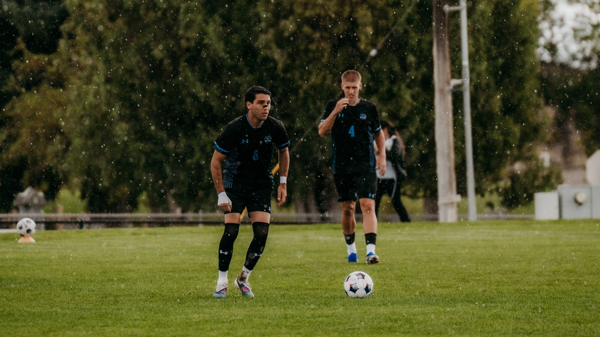 An action photograph of a Utah State Eastern men's soccer player controlling the ball during rainfall, with visible rain droplets falling over the grass pitch. A teammate in a dark navy jersey (number 4) follows closely behind him, set against a backdrop of green trees and utility poles.