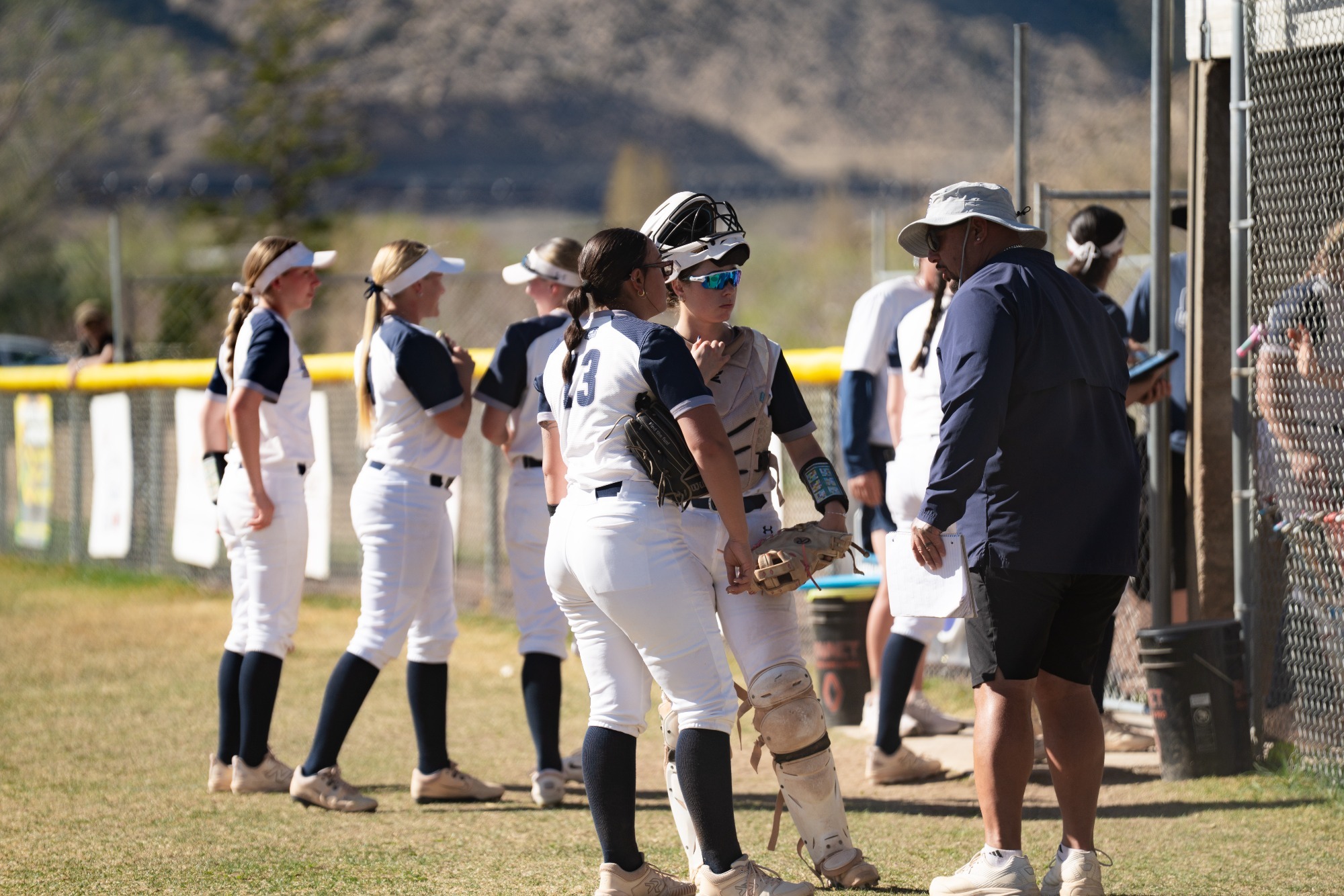 Softball players in white uniforms gather near the dugout as a catcher in gear speaks with a coach holding a clipboard.