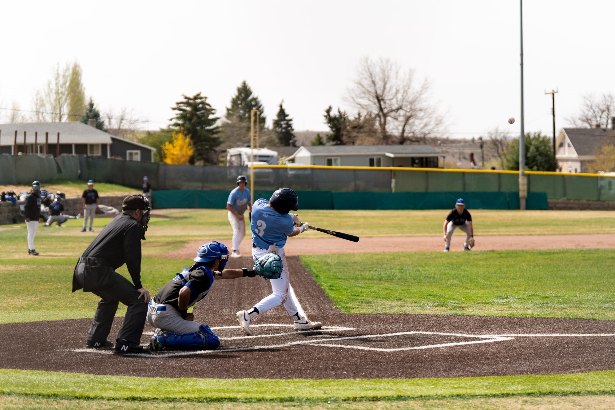 Baseball batter in a light blue jersey swings and makes contact at home plate as the catcher and umpire watch, with the ball flying into the outfield.